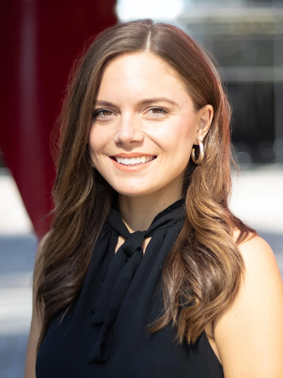 A smiling woman with long, wavy brown hair, wearing a black sleeveless top with a tie at the neckline and gold hoop earrings, outdoors with a blurred background.