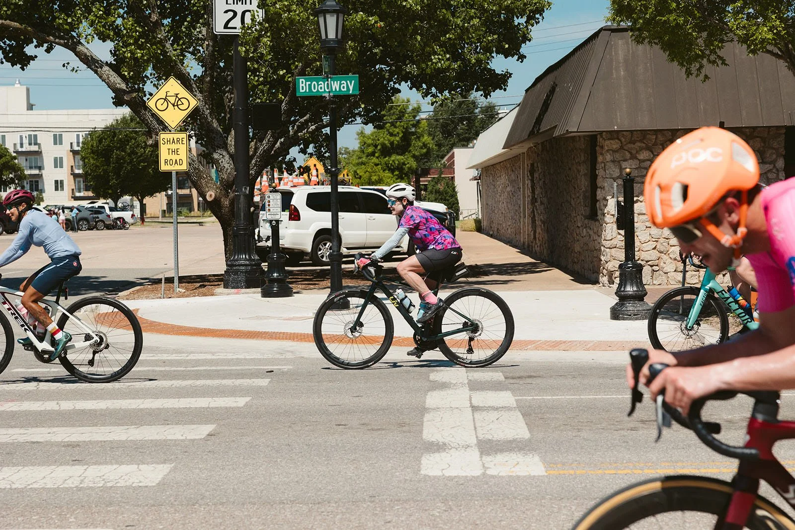 People riding bicycles across a city street at a crosswalk on a sunny day, with trees, parked cars, and buildings in the background.