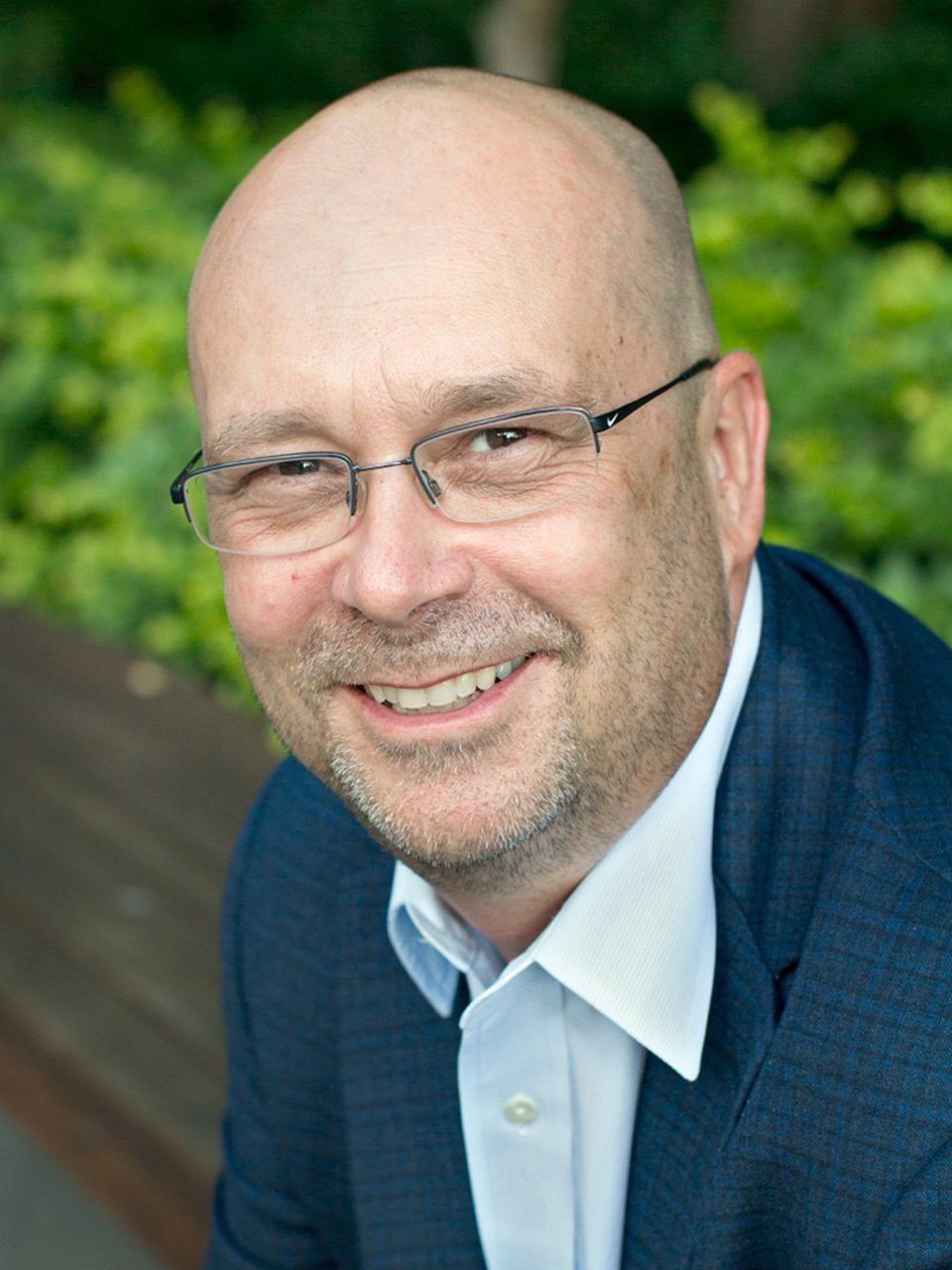 Close-up portrait of a smiling middle-aged man with glasses, a bald head, and a beard, wearing a blue jacket and white shirt, with a green leafy background.
