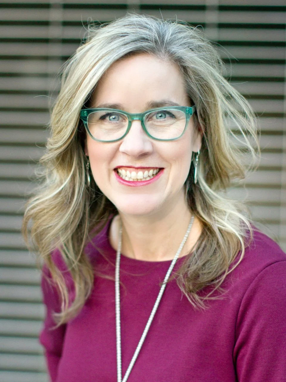 A woman with wavy blonde hair, wearing teal glasses, a maroon top, and a necklace, smiling outdoors with a wooden fence in the background.