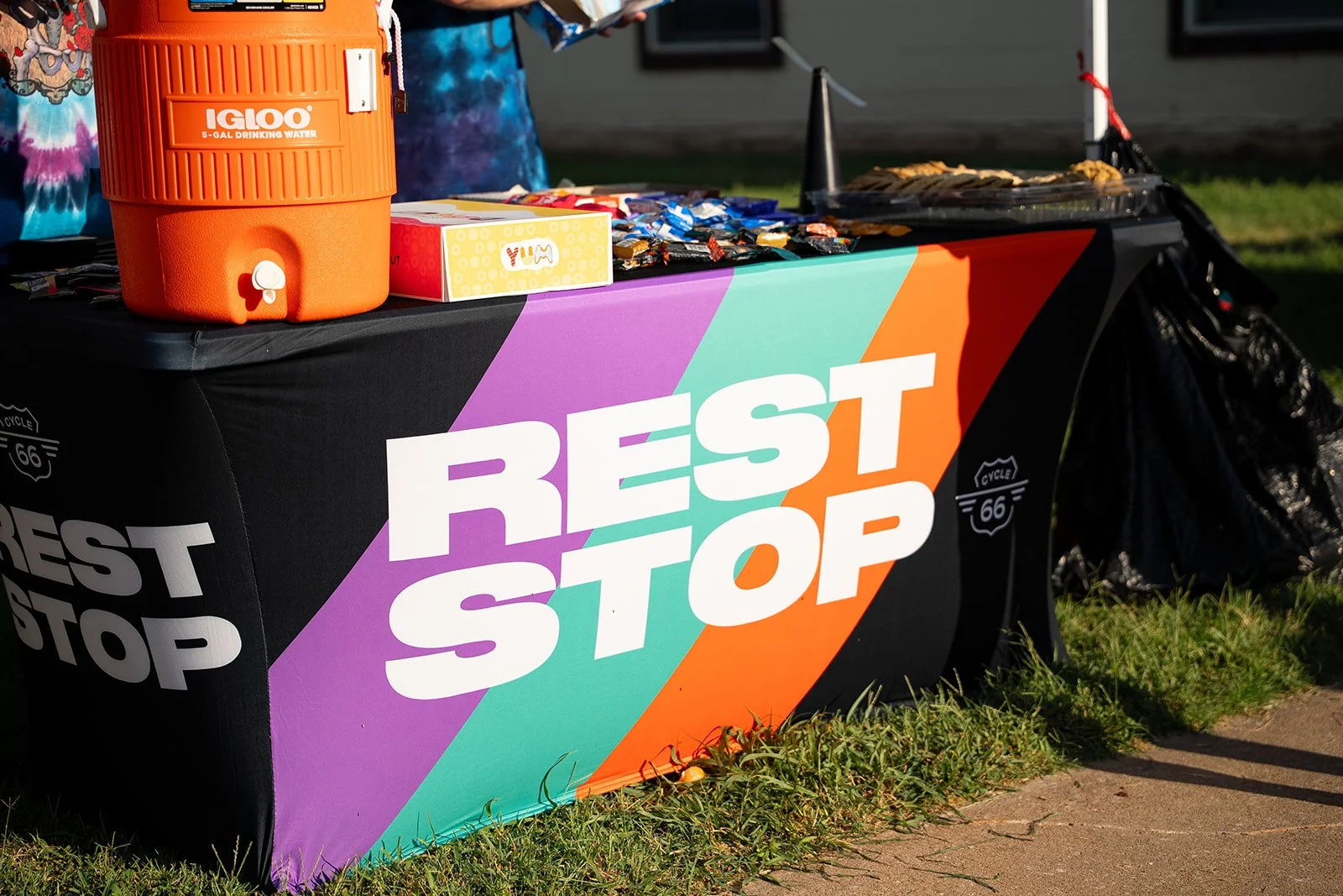 A table with a colorful tablecloth displaying snacks and drinks, with a large orange water cooler and a black trash bag nearby. The tablecloth has the words "REST STOP" printed on it.