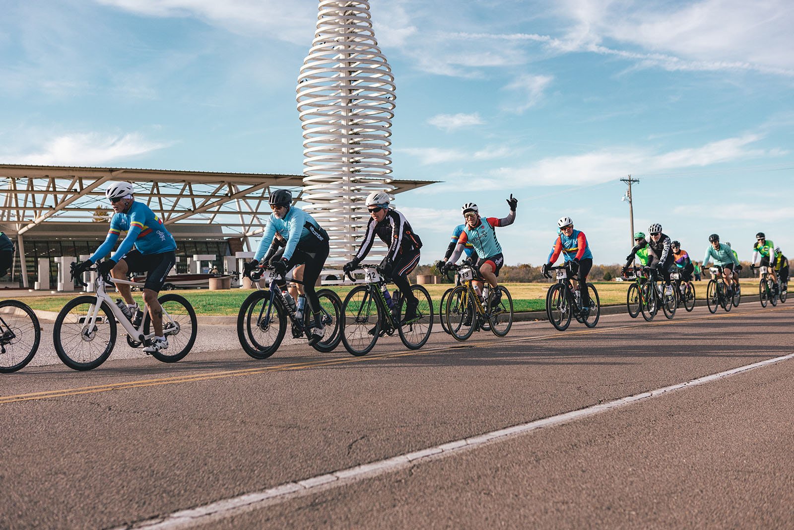 A group of cyclists riding on a paved road during daytime, with some wearing helmets and colorful jerseys, and one cyclist raising an arm. The background features a modern architectural structure and open fields, under a partly cloudy sky.