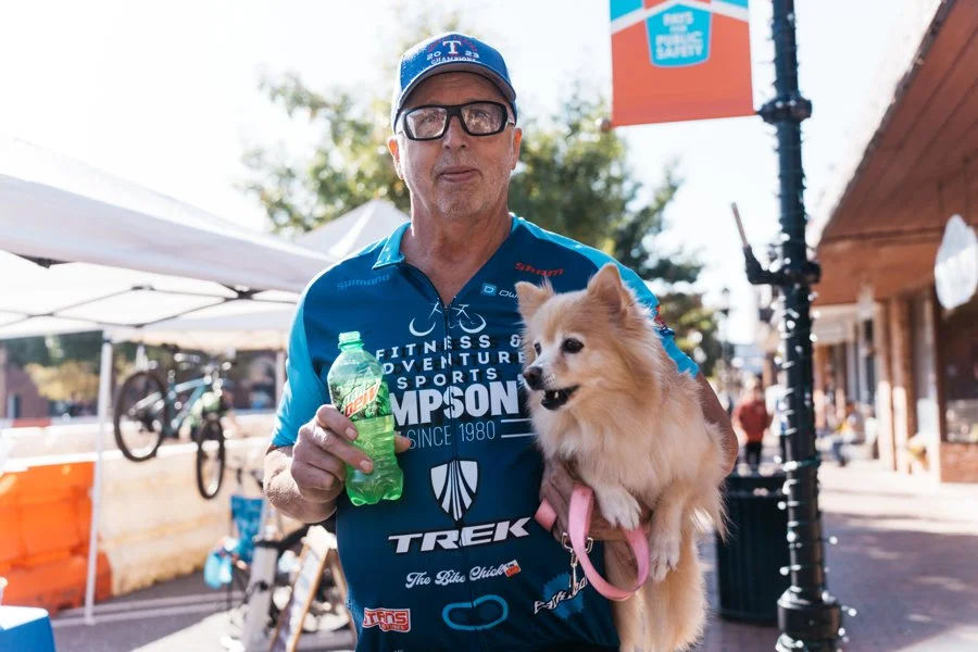 Older man wearing sunglasses and cycling jersey holding a bottle of Mountain Dew and a small dog on pink leash in an outdoor setting.