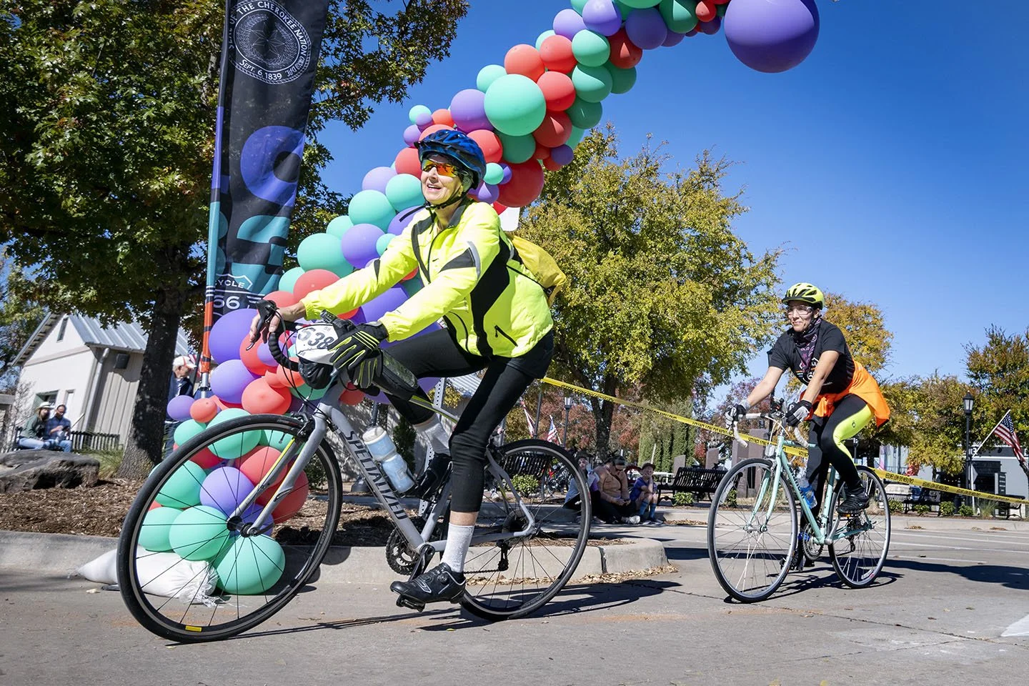 Two women riding bicycles in a park during a city cycling event, with colorful balloon decorations and spectators in the background.