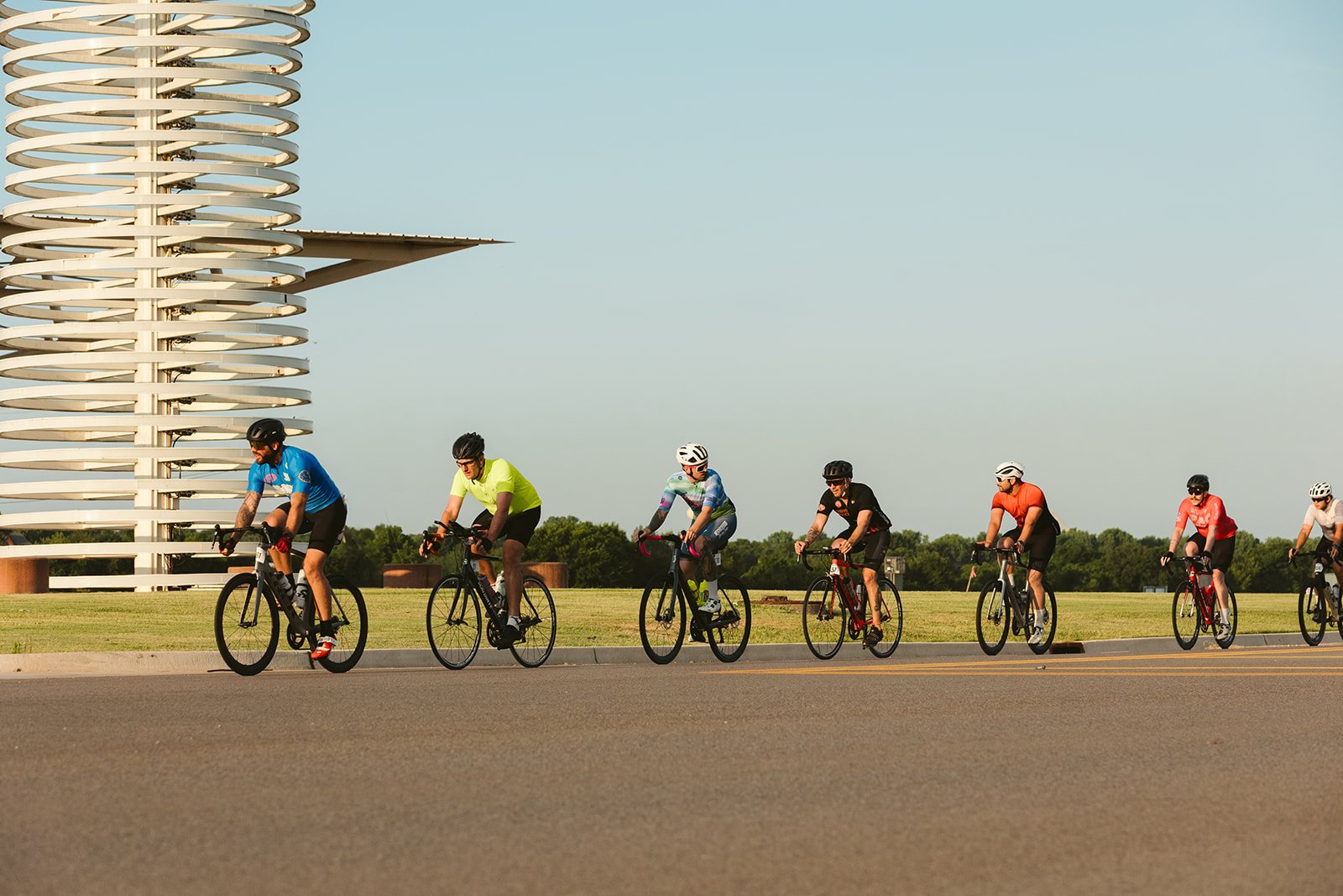 Group of seven cyclists riding on the road near a modern sculptural building, with trees in the background on a clear day.