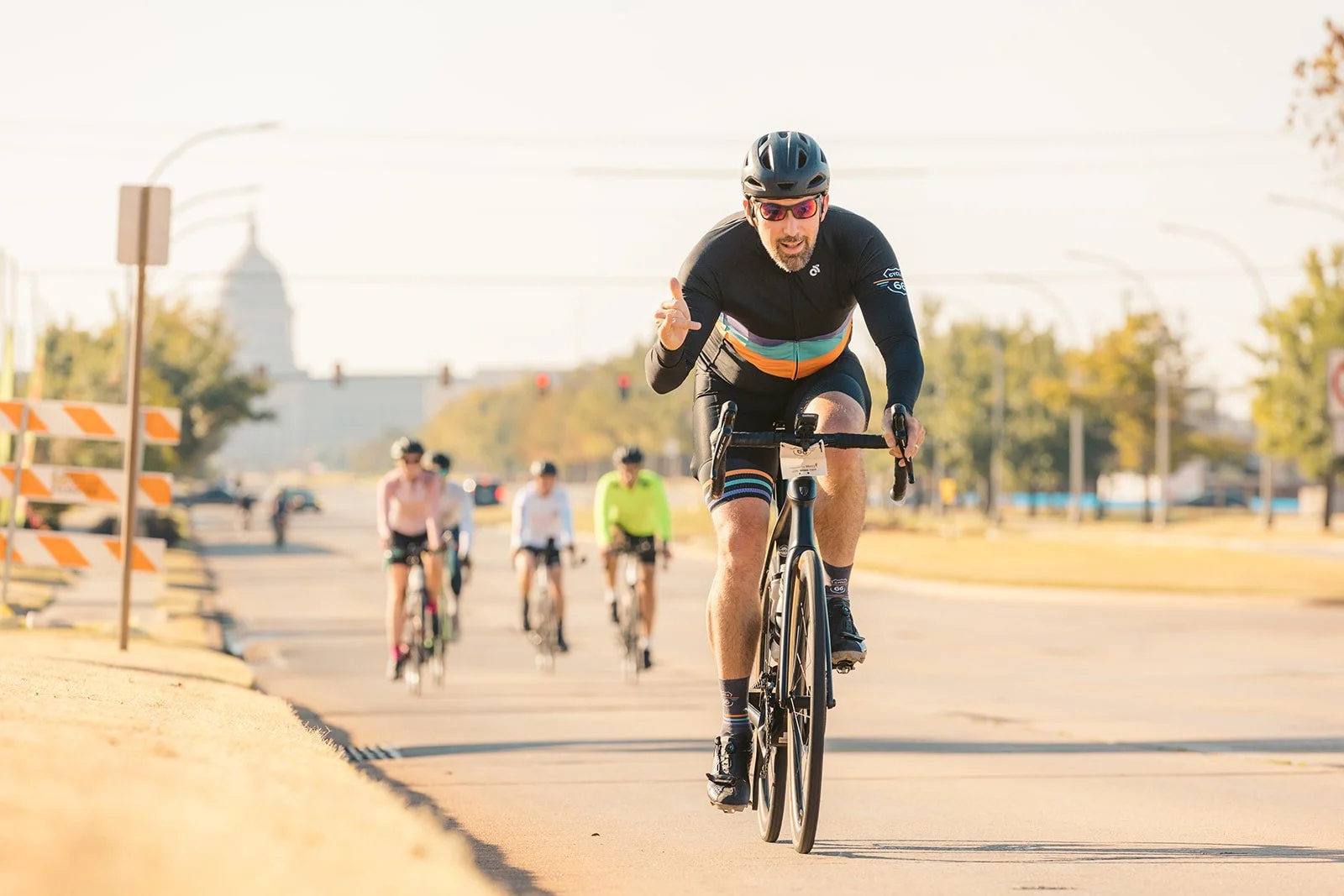 A man wearing a black cycling helmet, sunglasses, and a black long-sleeve cycling jersey with colorful stripes rides a bicycle on a paved path, with several other cyclists in the background.