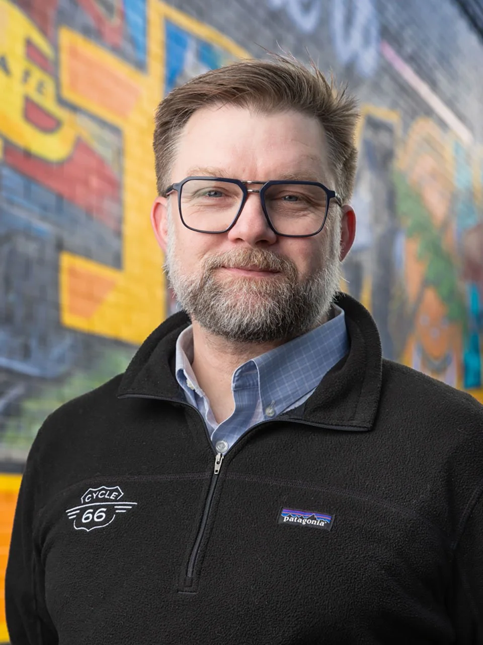 A man with glasses and a beard standing in front of a colorful mural.