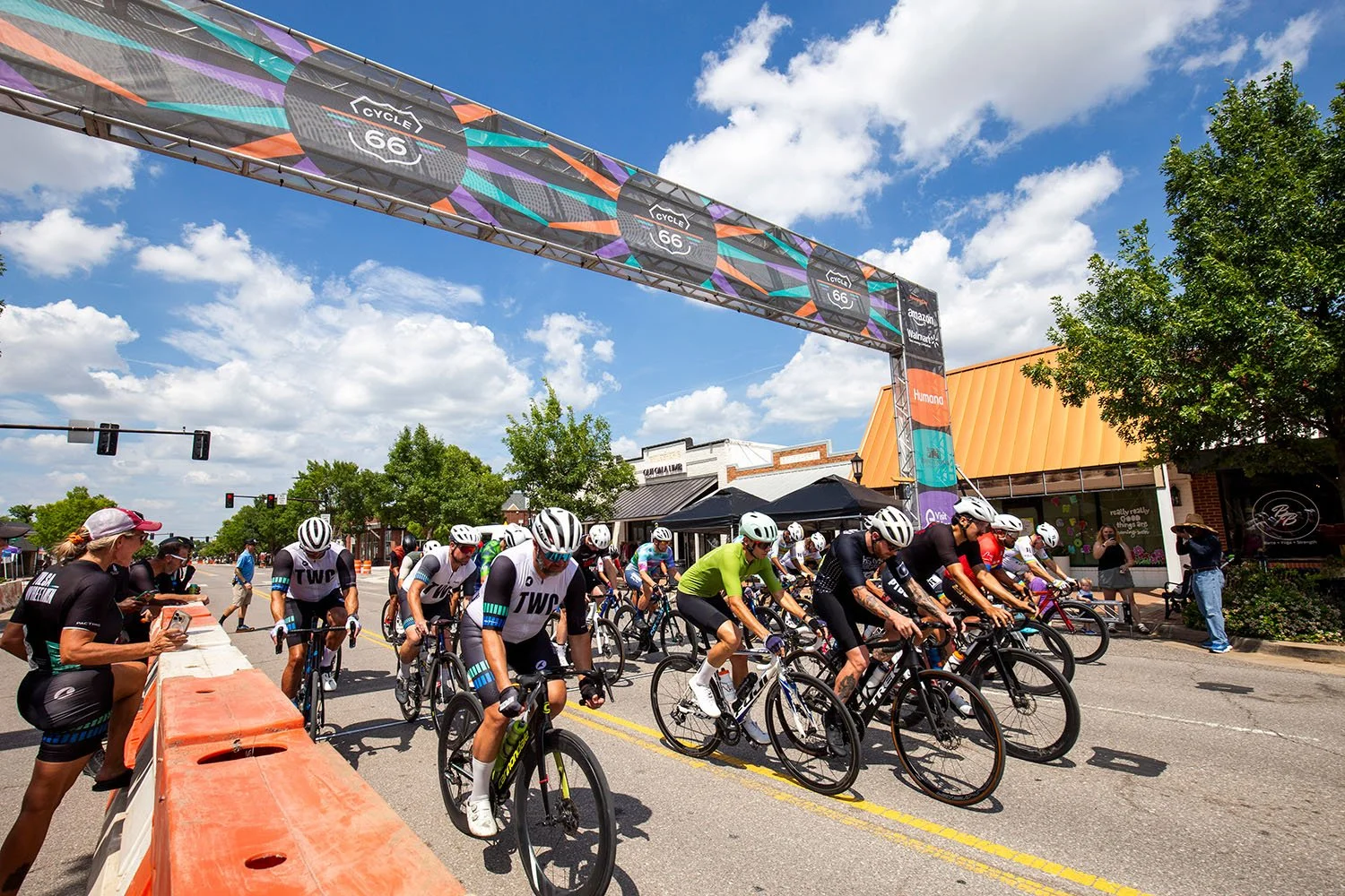 Bicycle race with cyclists starting at the starting line under a colorful banner reading "Cycle 66"; spectators watch on the sides on a sunny day with blue sky and white clouds.