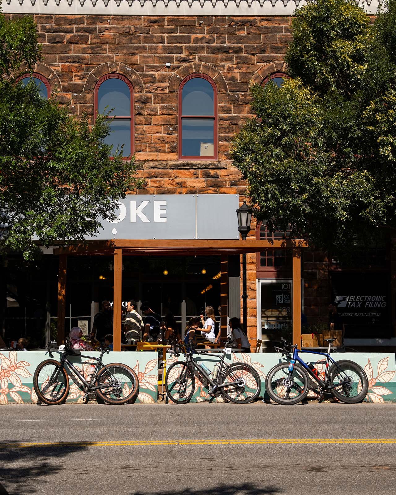 A row of three bicycles parked along the curb in front of a restaurant with an outdoor seating area. The restaurant has a brick facade with three arched windows on the upper level and a partial sign that reads 'OKE'. People are sitting and standing inside and outside the restaurant, with trees partially obscuring the view.