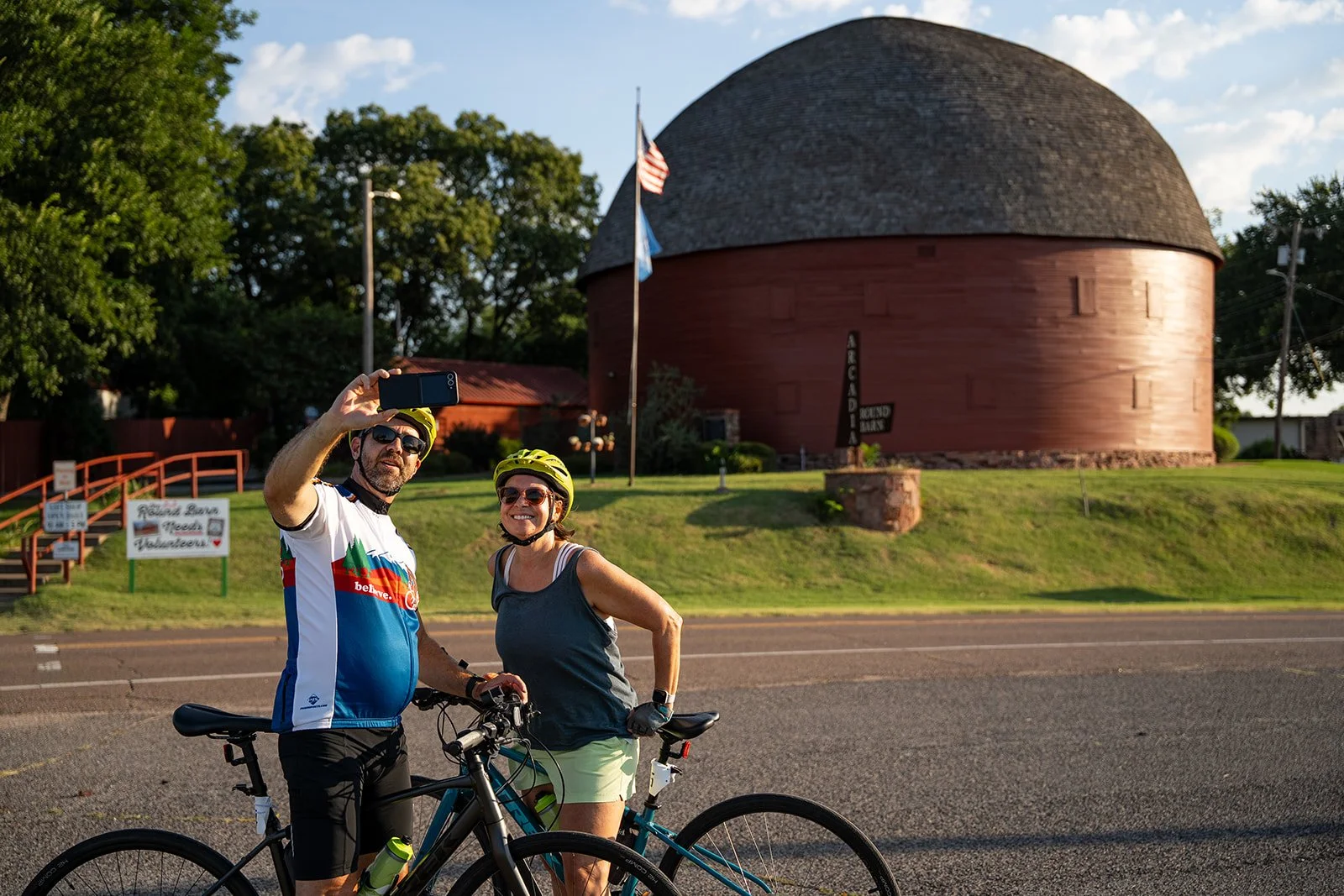 A man and woman in cycling gear taking a selfie with bicycles in front of a round red building with a sign that says 'Round Barn' and a flagpole with the American flag. The scene is outdoors, likely in the evening.
