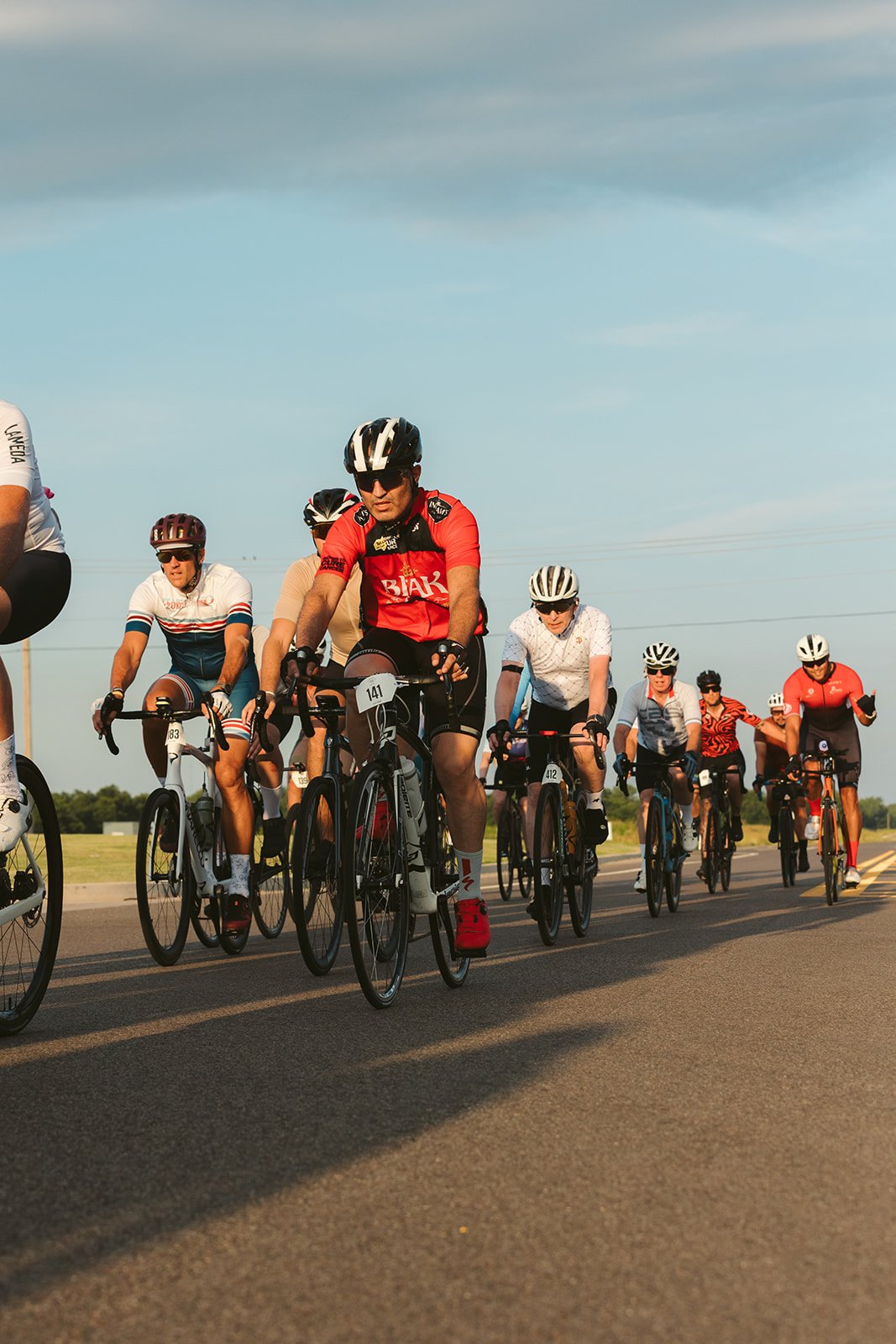 Group of people riding bicycles on a paved road under a blue sky, wearing helmets and athletic clothing.