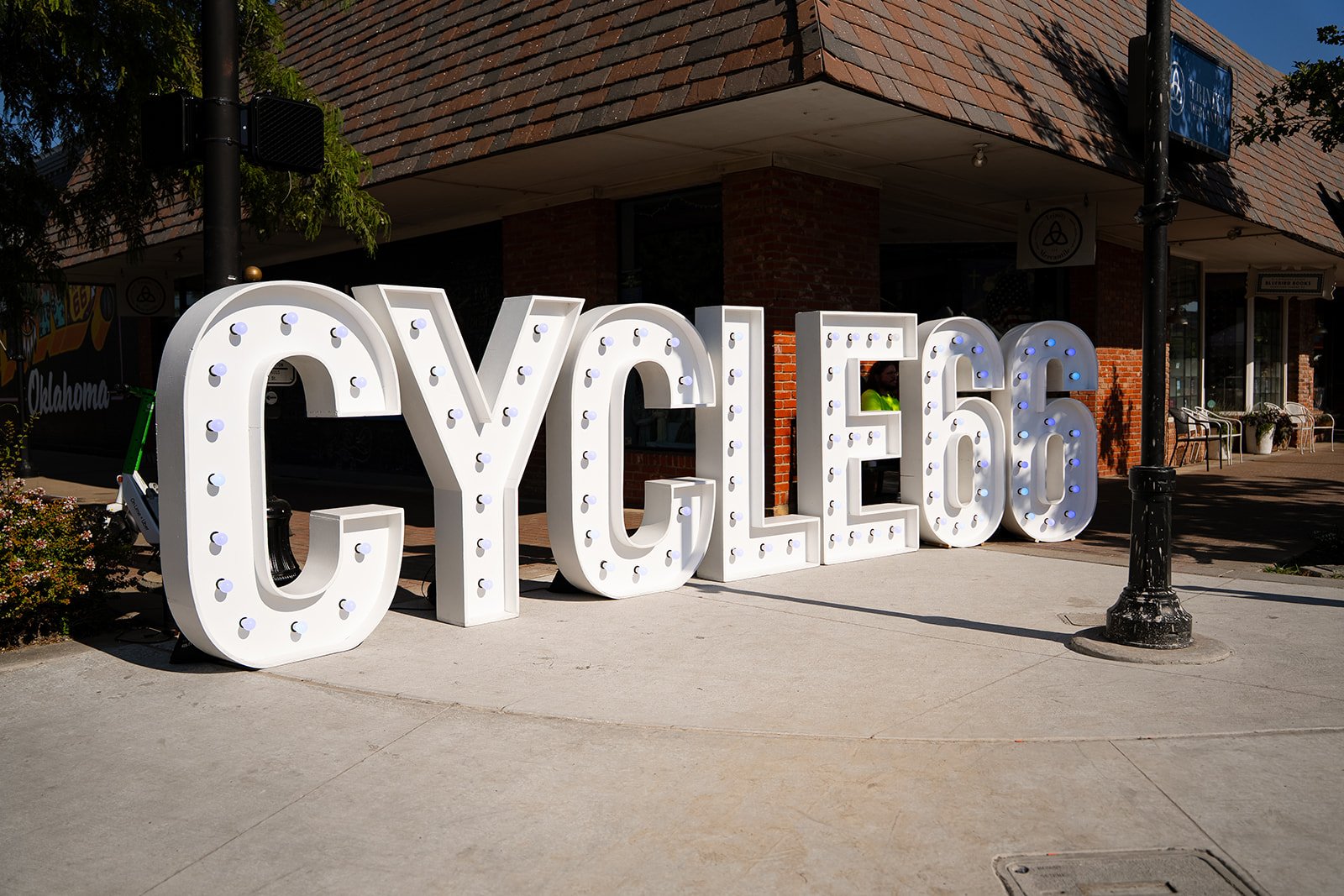 Large white illuminated letters spelling 'CYCLE 66' on the sidewalk in front of a red brick building, with a person in a yellow vest visible through a window.