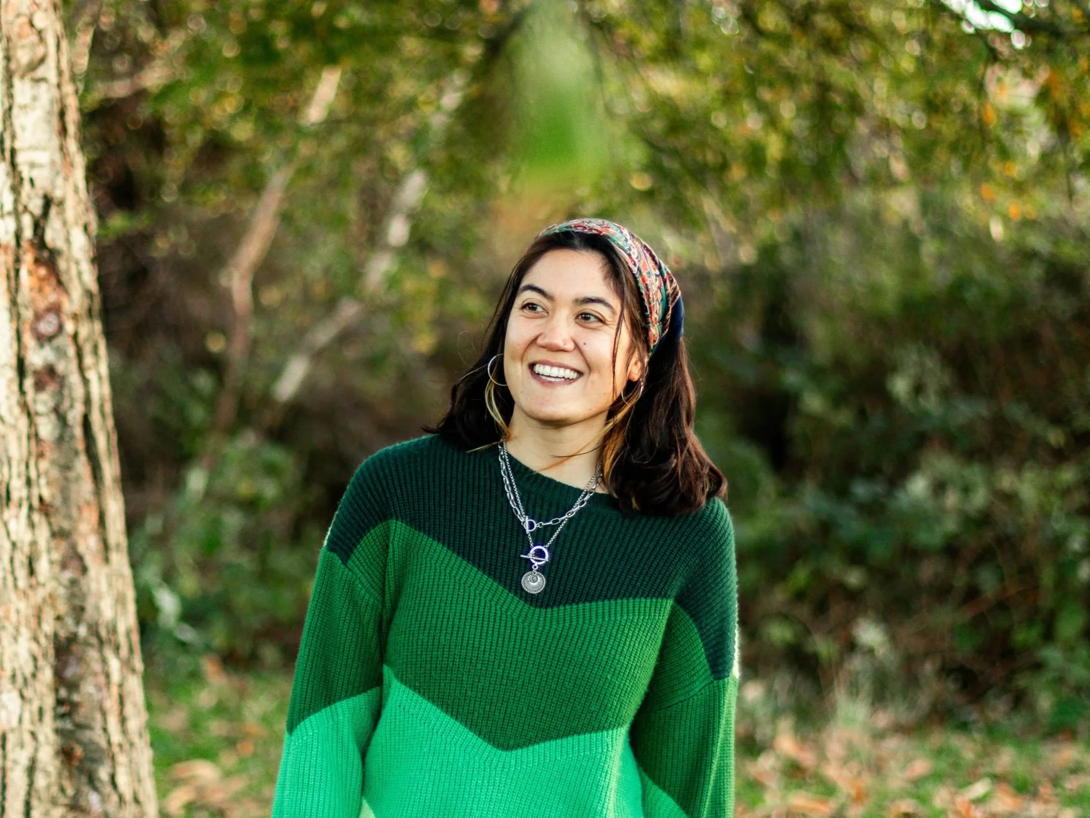 A woman with dark hair wearing a multicolored headscarf, hoop earrings, necklaces, and a green sweater is smiling outdoors in a forested area with trees and greenery.