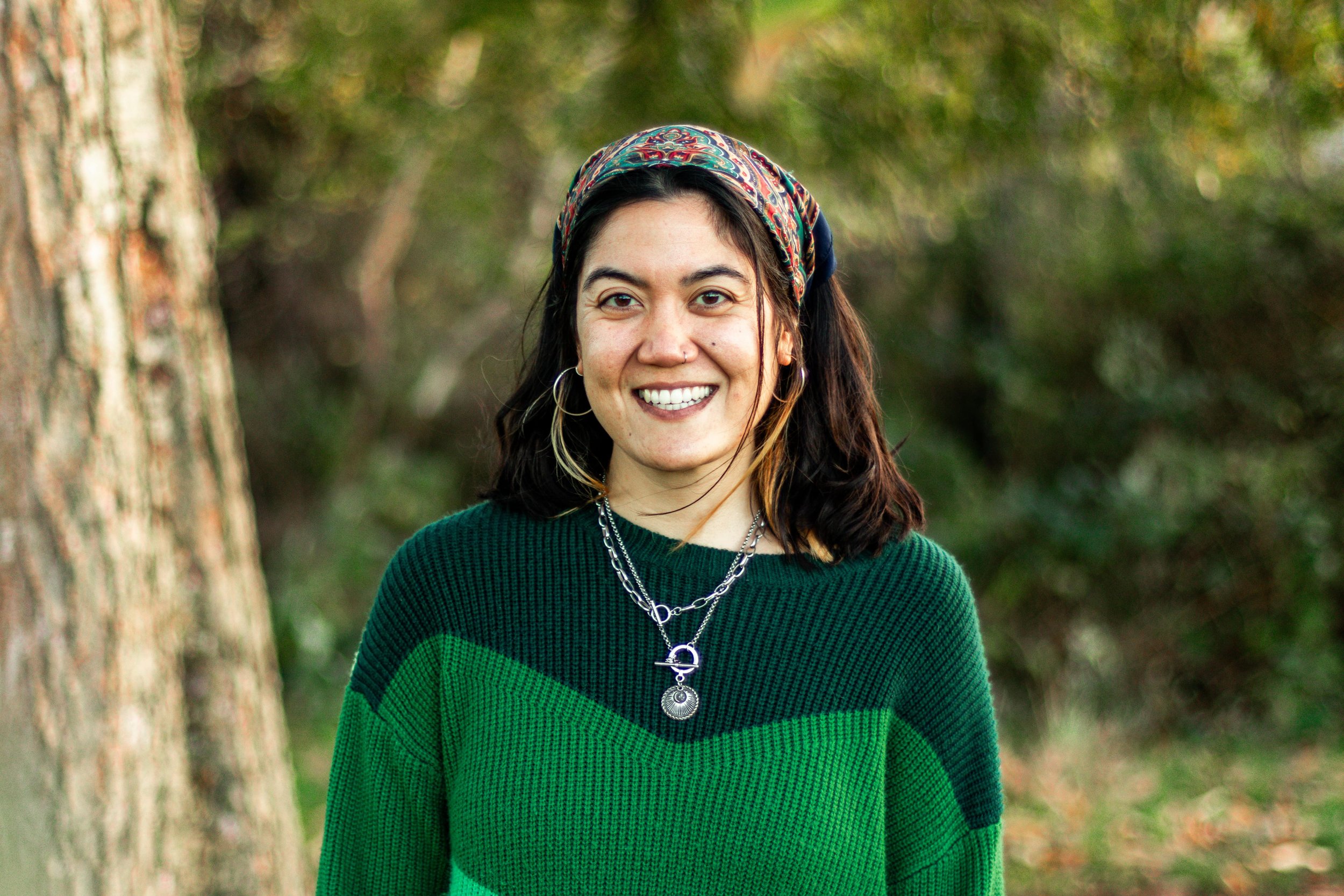 A woman outdoors smiling, wearing a green sweater with dark green and light green sections, layered necklaces, hoop earrings, and a patterned headscarf, with trees and greenery in the background.