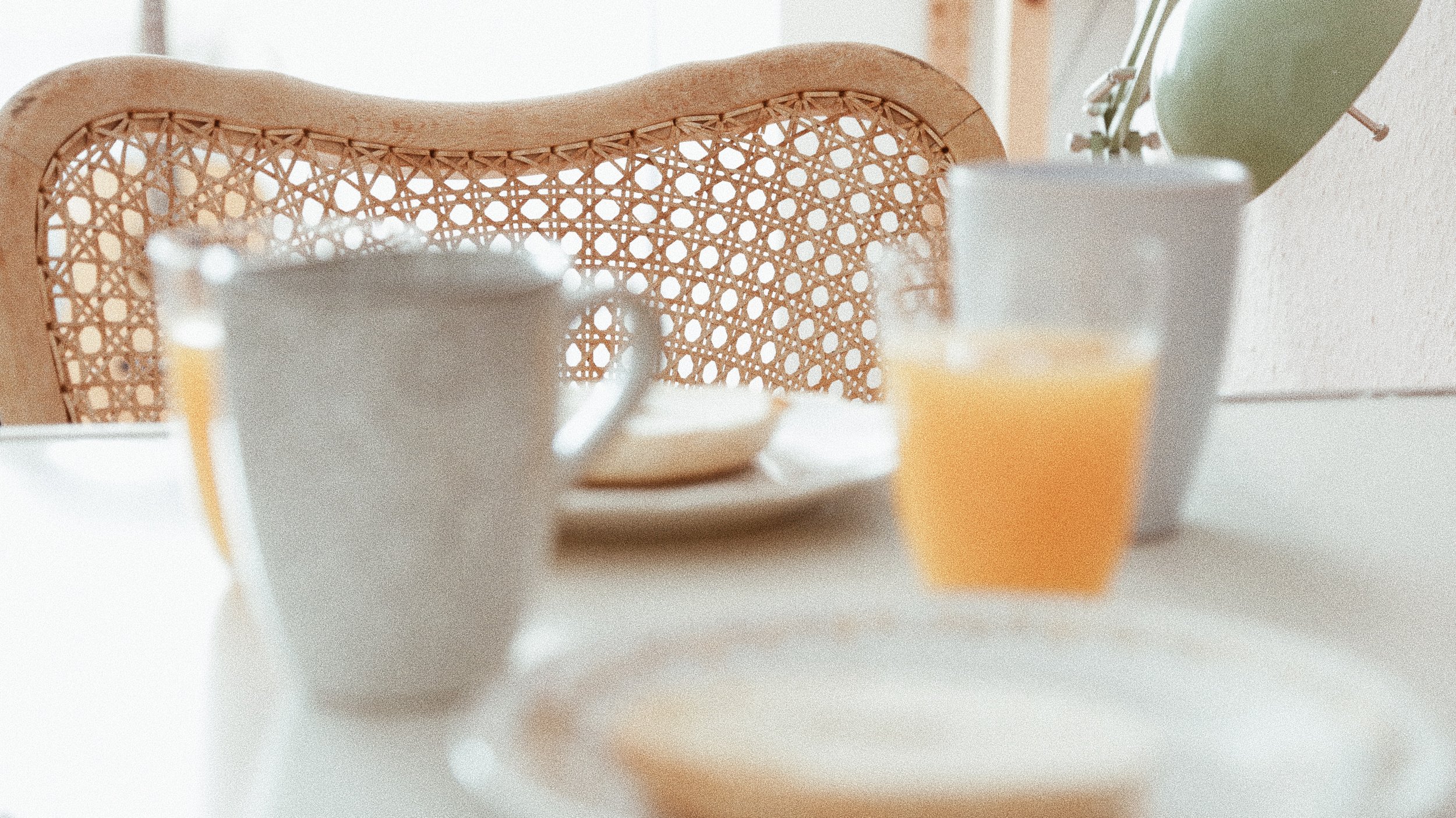 Frühstückstisch mit einer Tasse, einem Glas Orangensaft, einem kleinen Teller und einem weißen Topf mit einer Pflanze. Im Hintergrund ist ein Rattanstuhl.