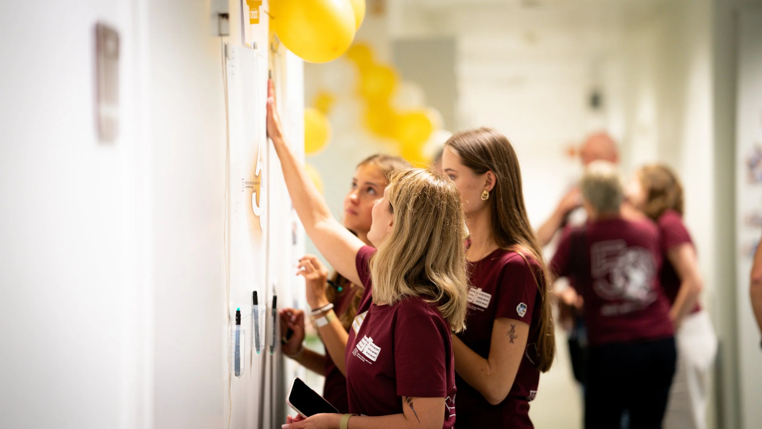 Mehrere Menschen, hauptsächlich Frauen, arbeiten an einer Pinnwand mit Notizen und Dokumenten, während sie an einer Veranstaltung oder Konferenz teilnehmen. Es sind gelbe Luftballons im Hintergrund sichtbar.