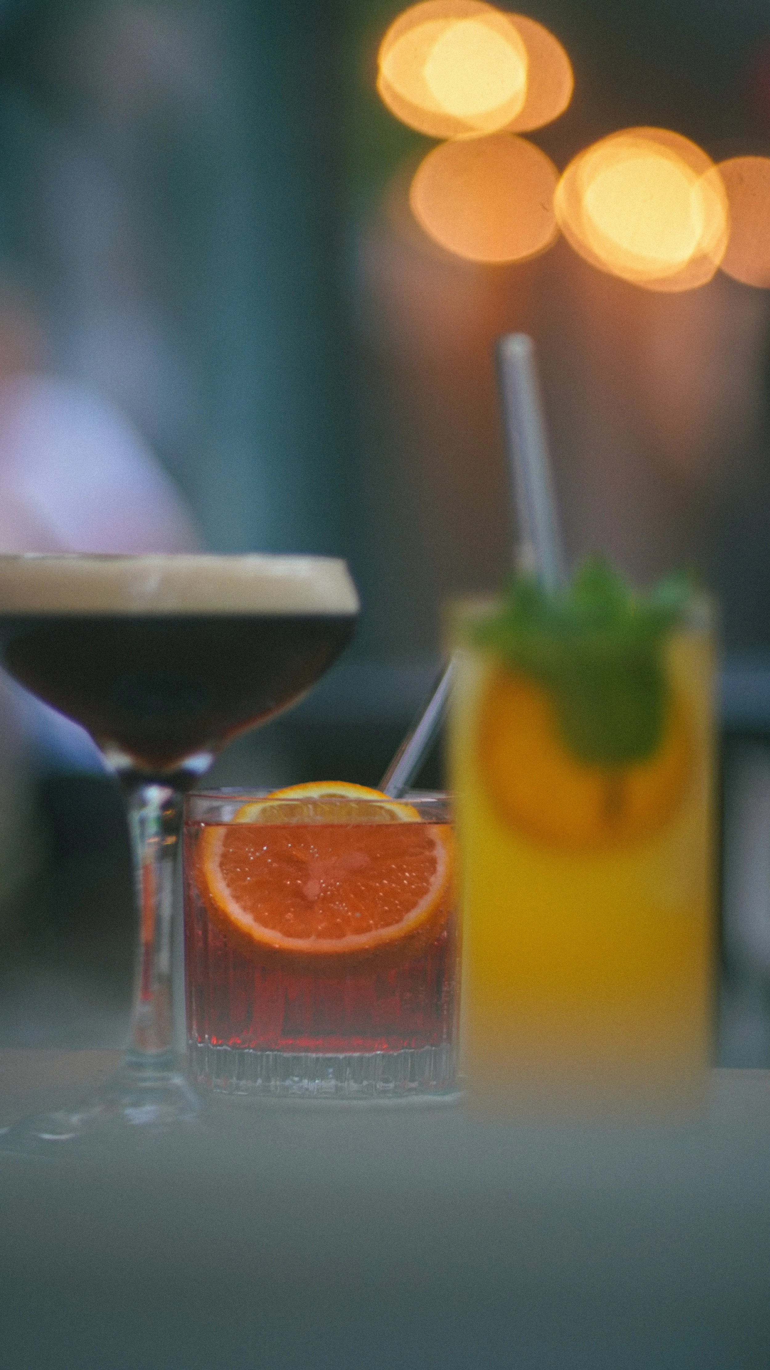 Drei bunte Cocktails mit Zitrusfrüchten auf einem Tisch, unscharfe Lichter im Hintergrund.