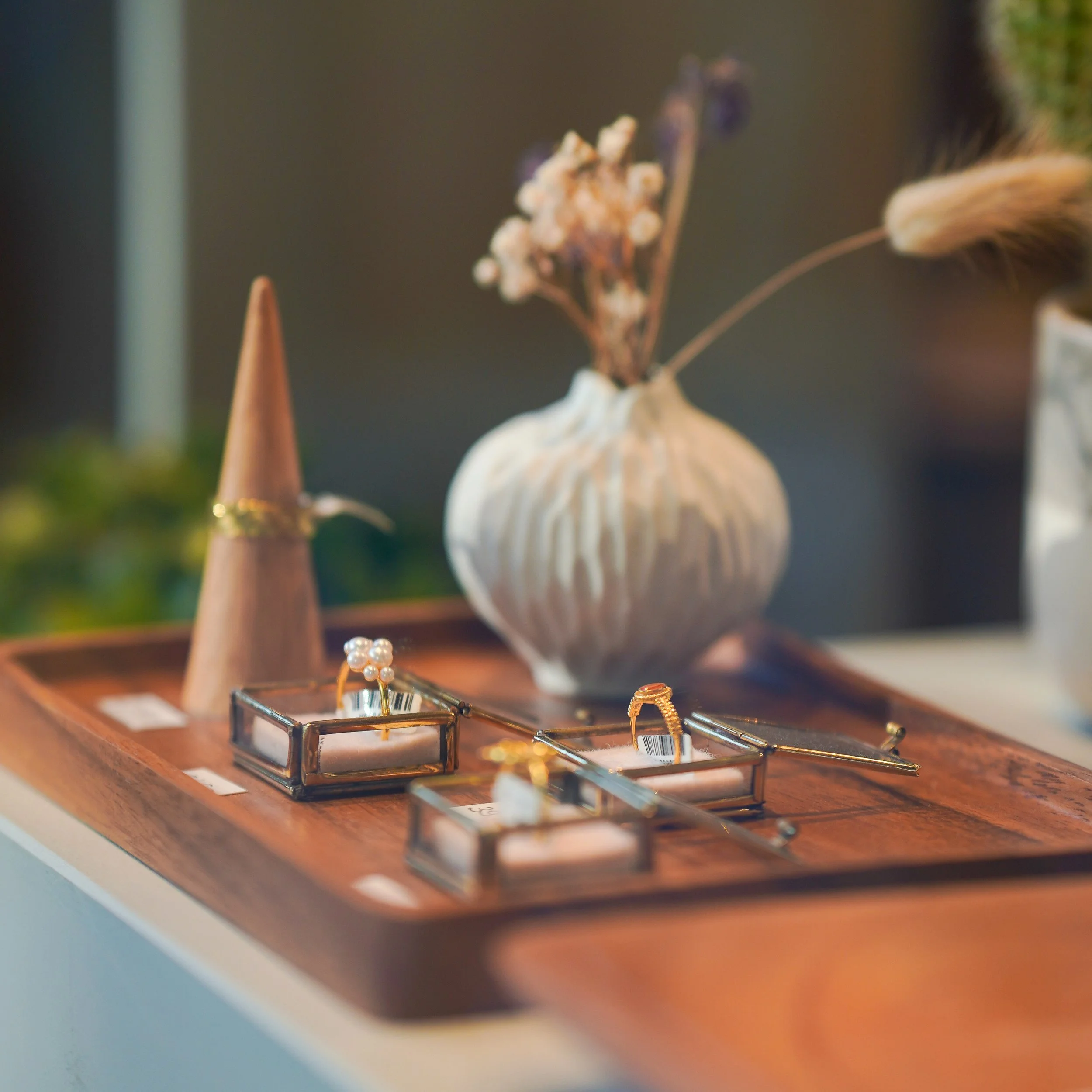 Ohrringe und Ringe auf einem Holztisch, im Hintergrund eine weiße Keramikvase mit getrockneten Blumen.