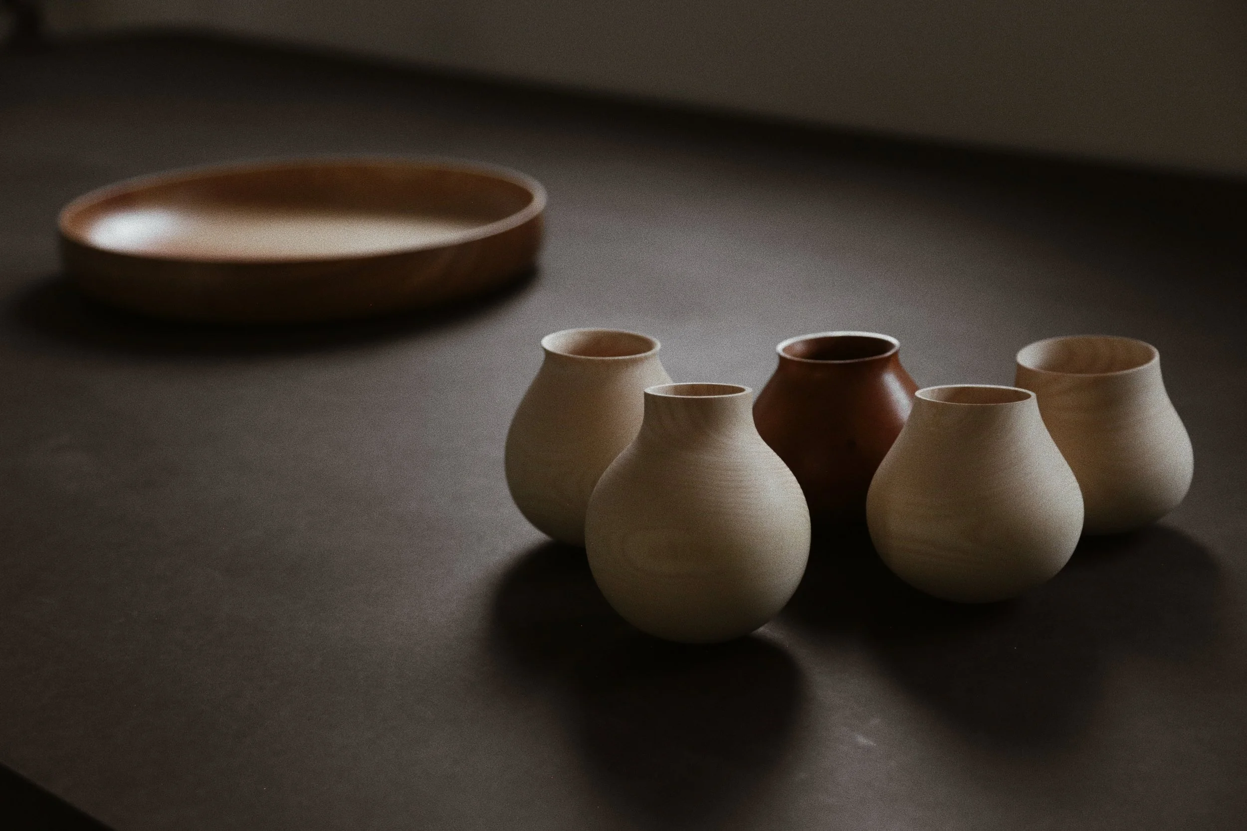 Fünf kleine Tonvasen in hellen und dunklen Farben auf dunklem Untergrund, im Hintergrund eine größere flache Schale.