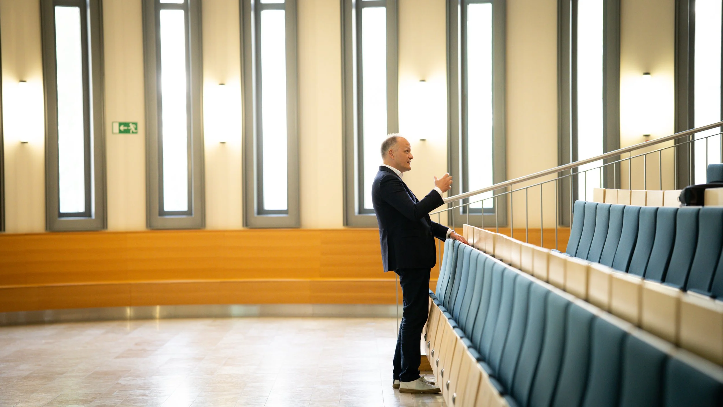 Ein Mann in Anzug steht in einem leeren, lichtdurchfluteten Saal vor leeren blauen Sitzreihen und blickt nach rechts.