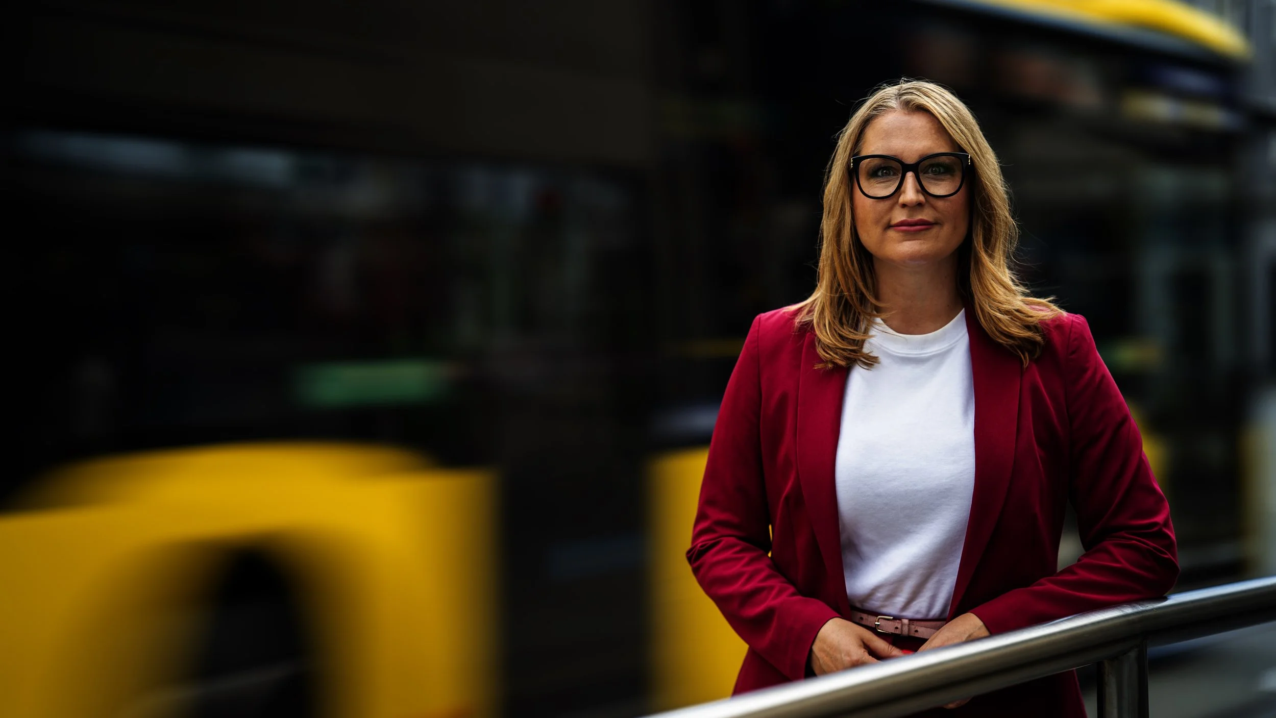 Eine Frau mit Brille und blonden Haaren trägt eine rosa Jacke und eine weiße Bluse und steht an einer Bushaltestelle in einer Stadt im Freien.