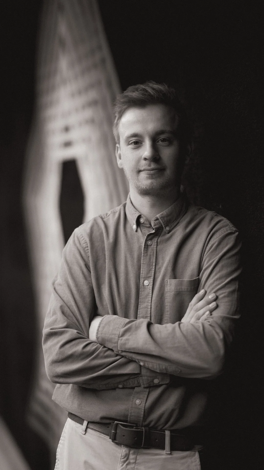 Schwarz-weiß-Fotografie eines jungen Mannes, der vor einer dunklen Wand steht, mit Armen verschränkt und einen leichten Lächeln zeigt.