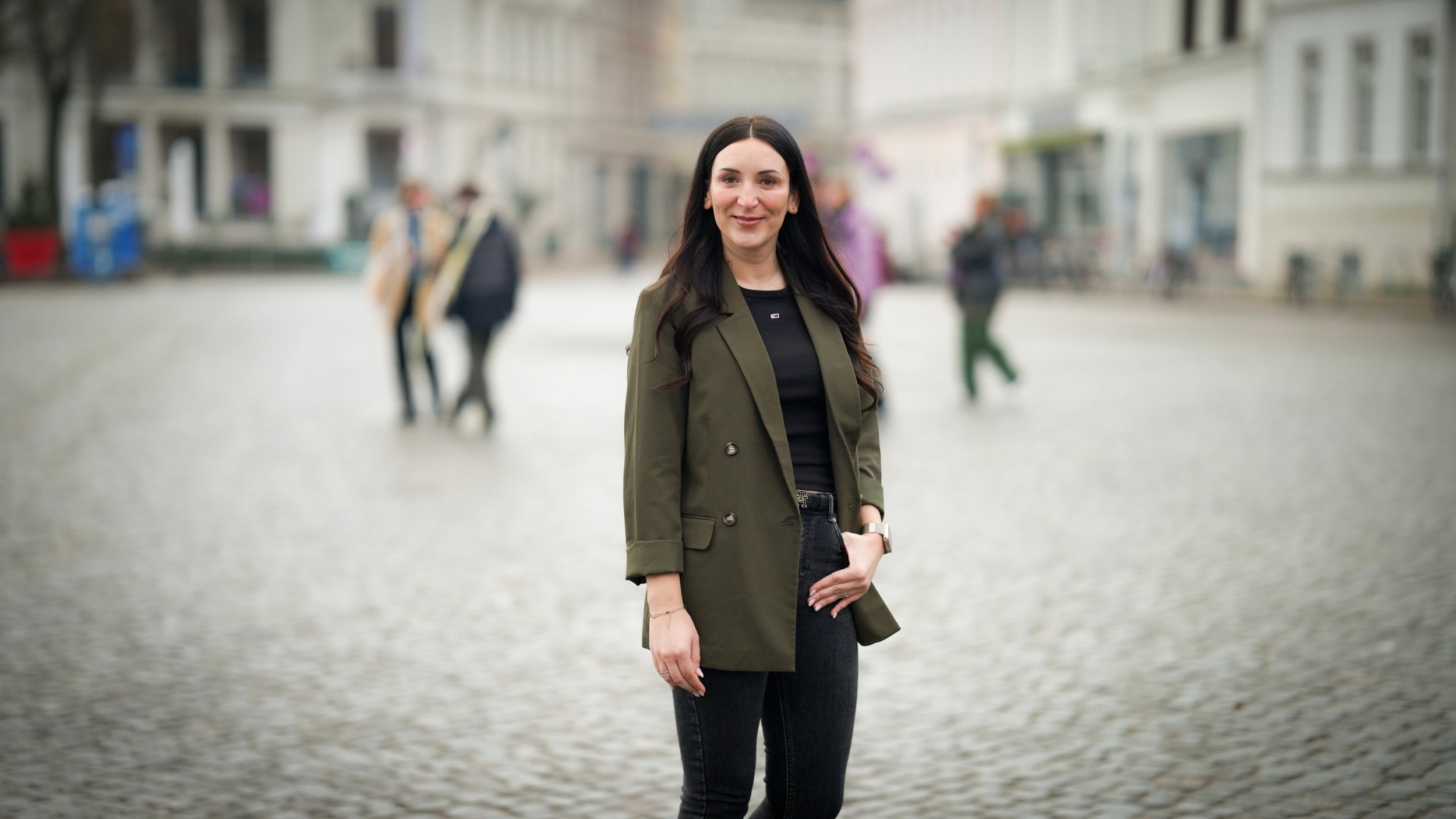 Eine junge Frau mit langen dunklen Haaren, die in einer Stadt auf einer gepflasterten Straße steht, trägt eine grünliche Jacke und schwarze Kleidung, mit mehreren Menschen im Hintergrund.