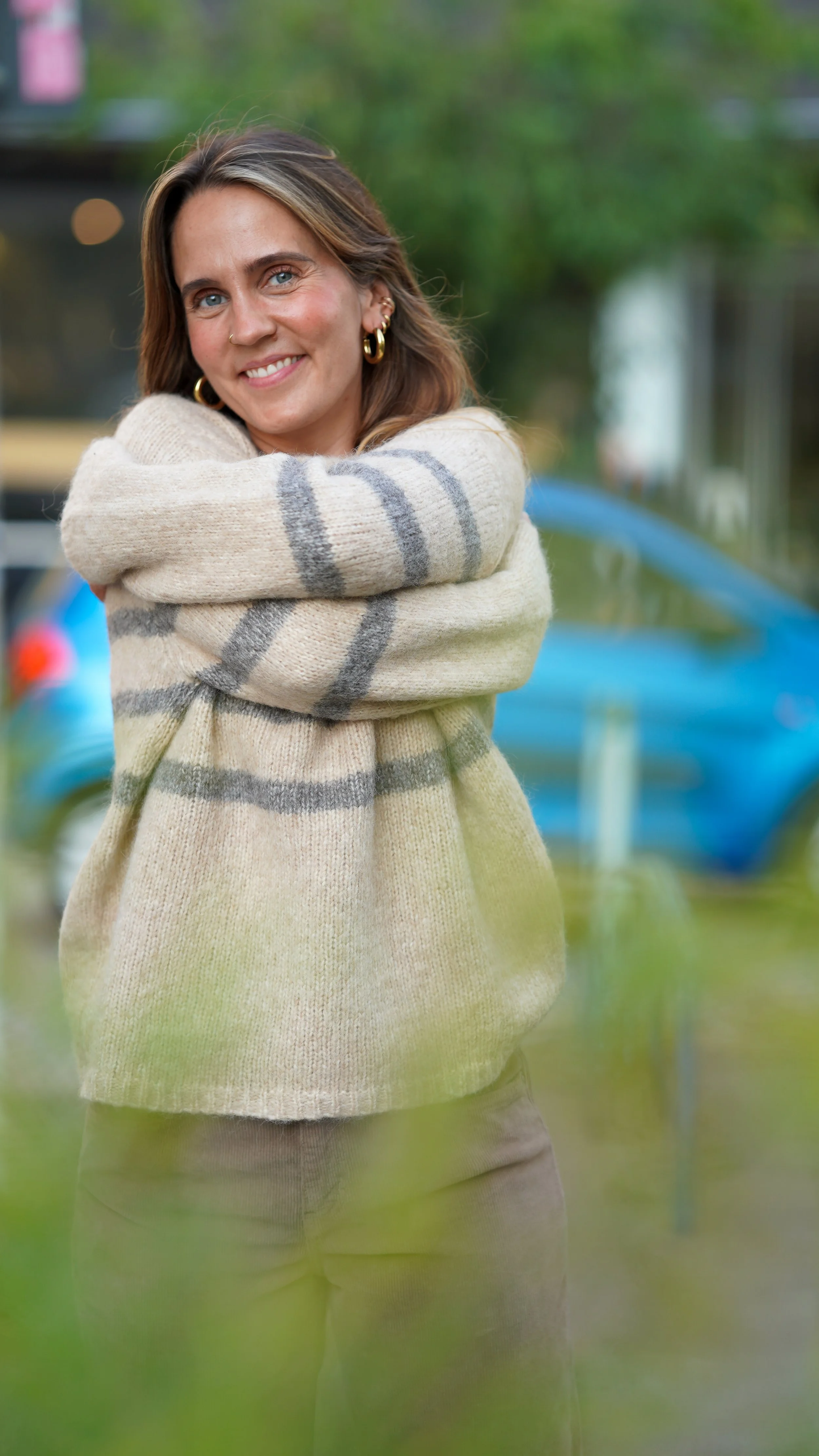 Eine lachende Frau in einem cremefarbenen Pullover mit grauen Streifen steht draußen vor einem blauen Auto, umgeben von grünen Bäumen.
