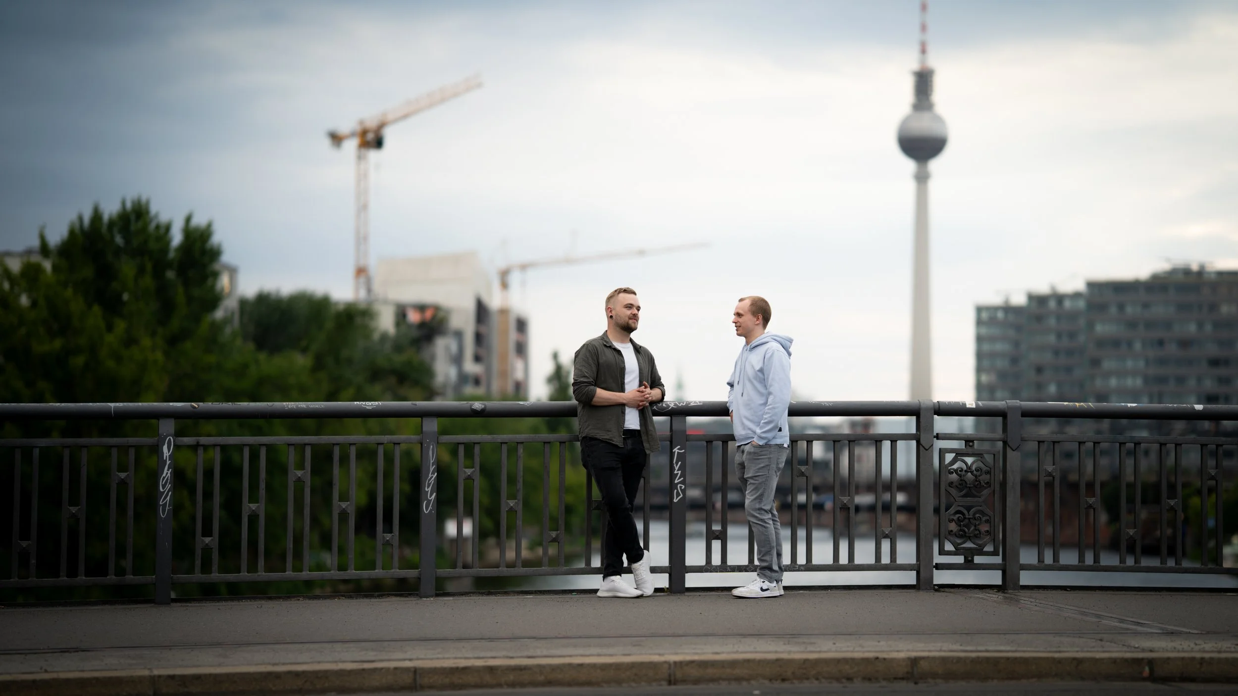 Zwei junge Männer stehen auf einer Brücke in Berlin, im Hintergrund der Fernsehturm und Baukräne, bei bedecktem Himmel.