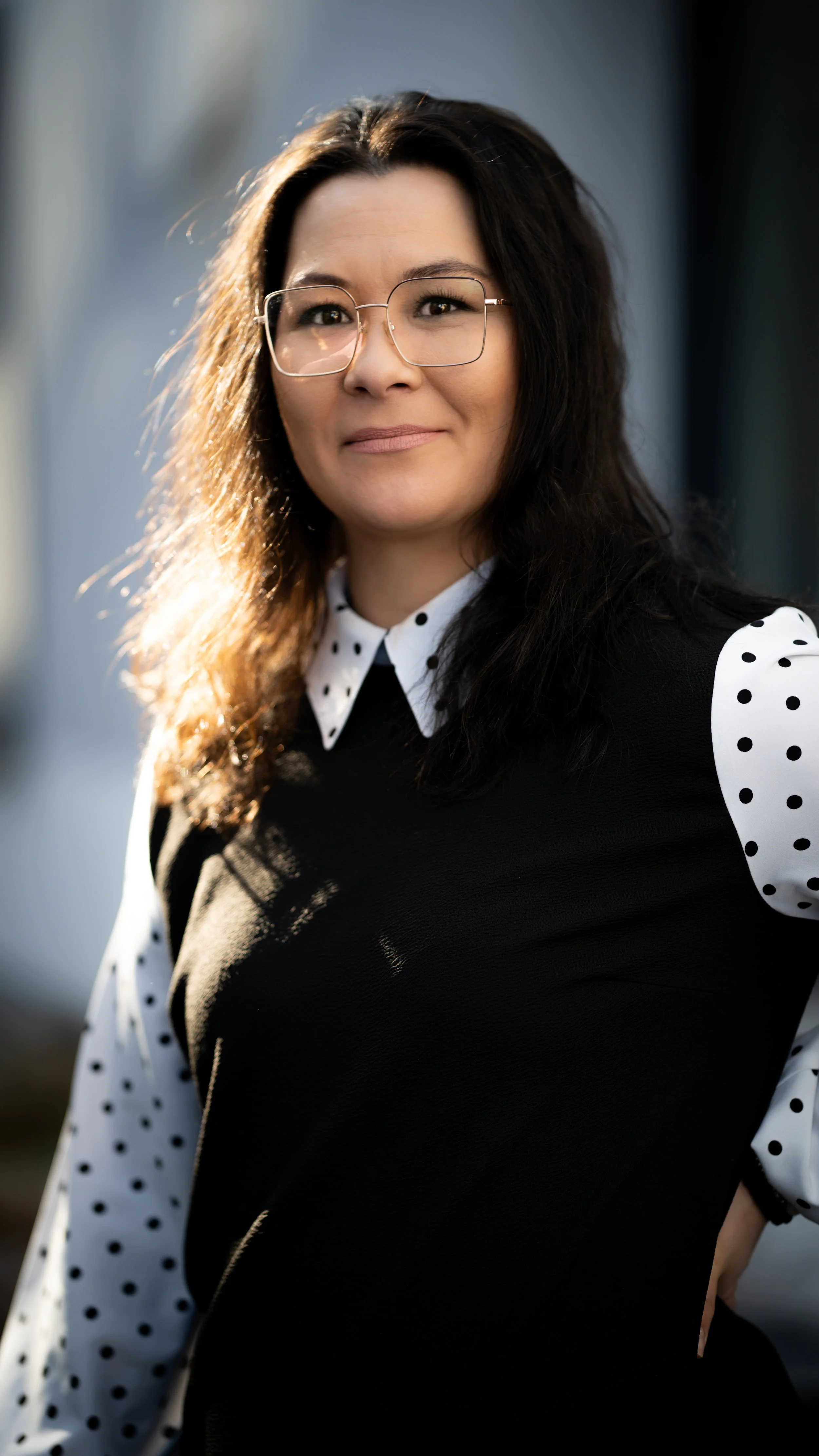 Frau mit Brille, schwarzem Pullover mit weißen, gepunkteten Ärmeln, steht im Abendlicht, lächelt leicht, Hintergrund unscharf.