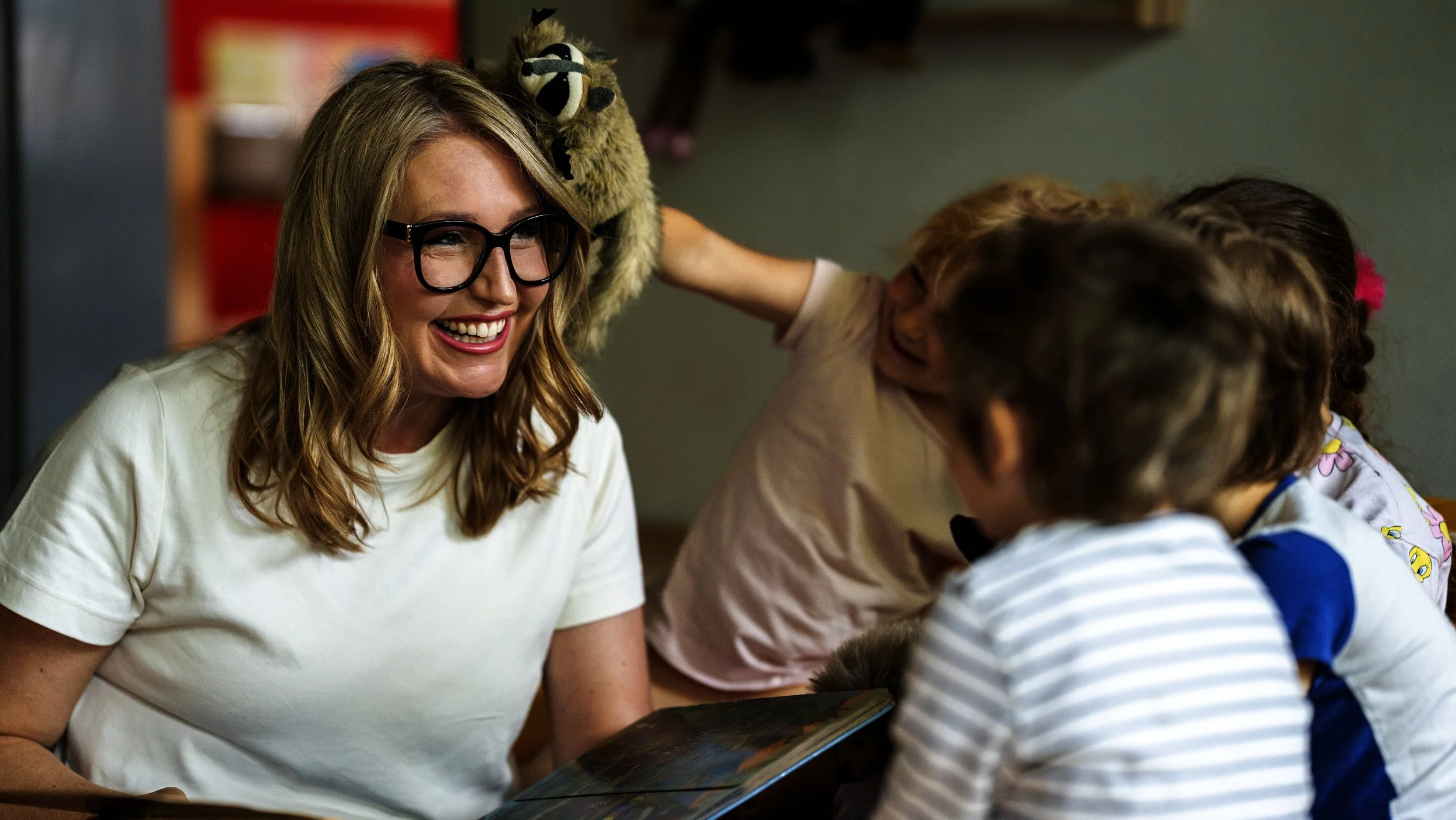 eine Frau mit Brille, die mit vier Kindern in einem Klassenzimmer lacht, während eines der Kinder ein Plüschtier auf ihren Kopf setzt