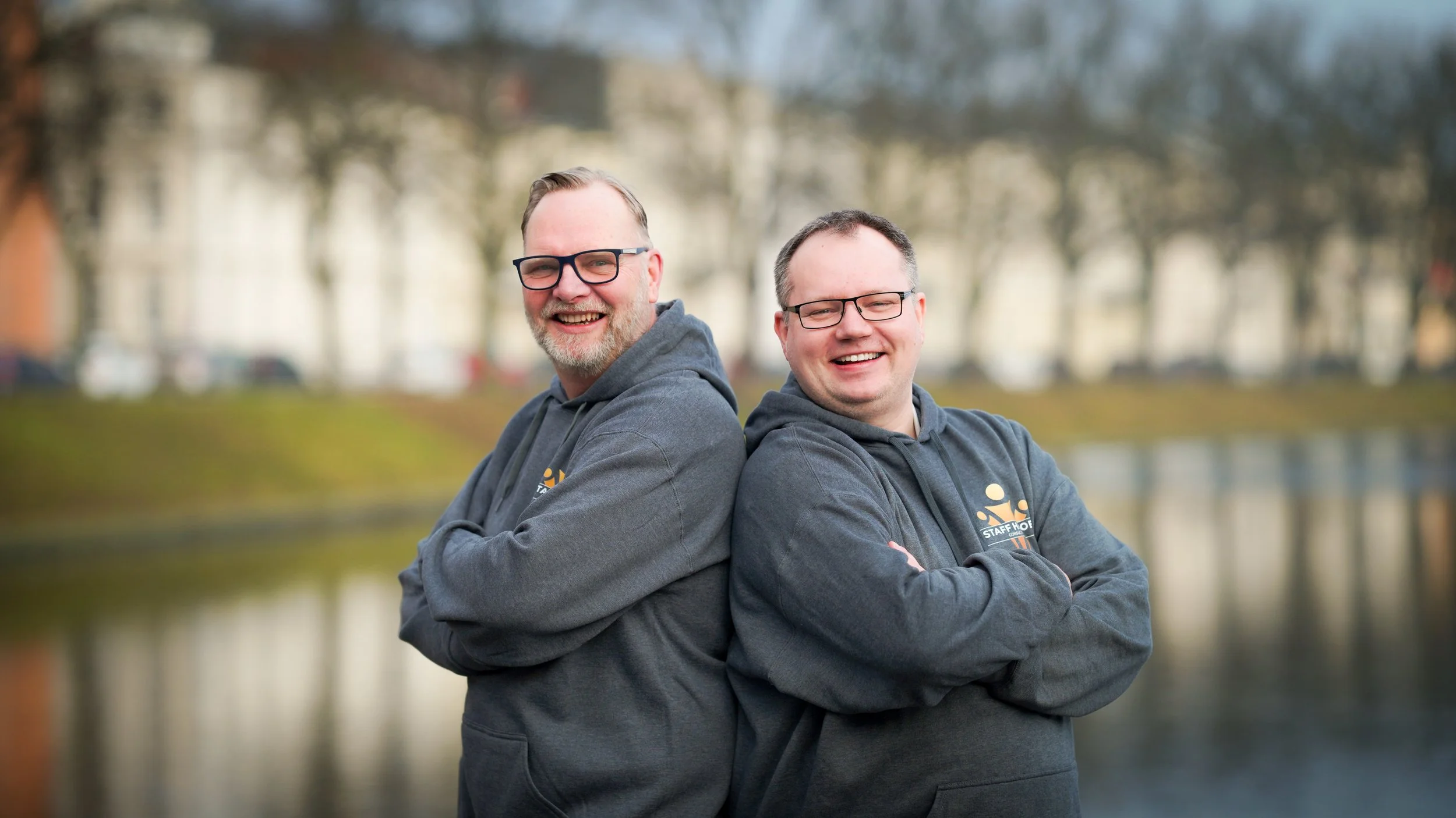 Zwei lachende Männer mit Brillen stehen Rücken an Rücken vor einer Wasserlandschaft, tragen dunkle Kapuzenpullover, im Hintergrund Bäume und Gebäude, an einem bewölkten Tag.