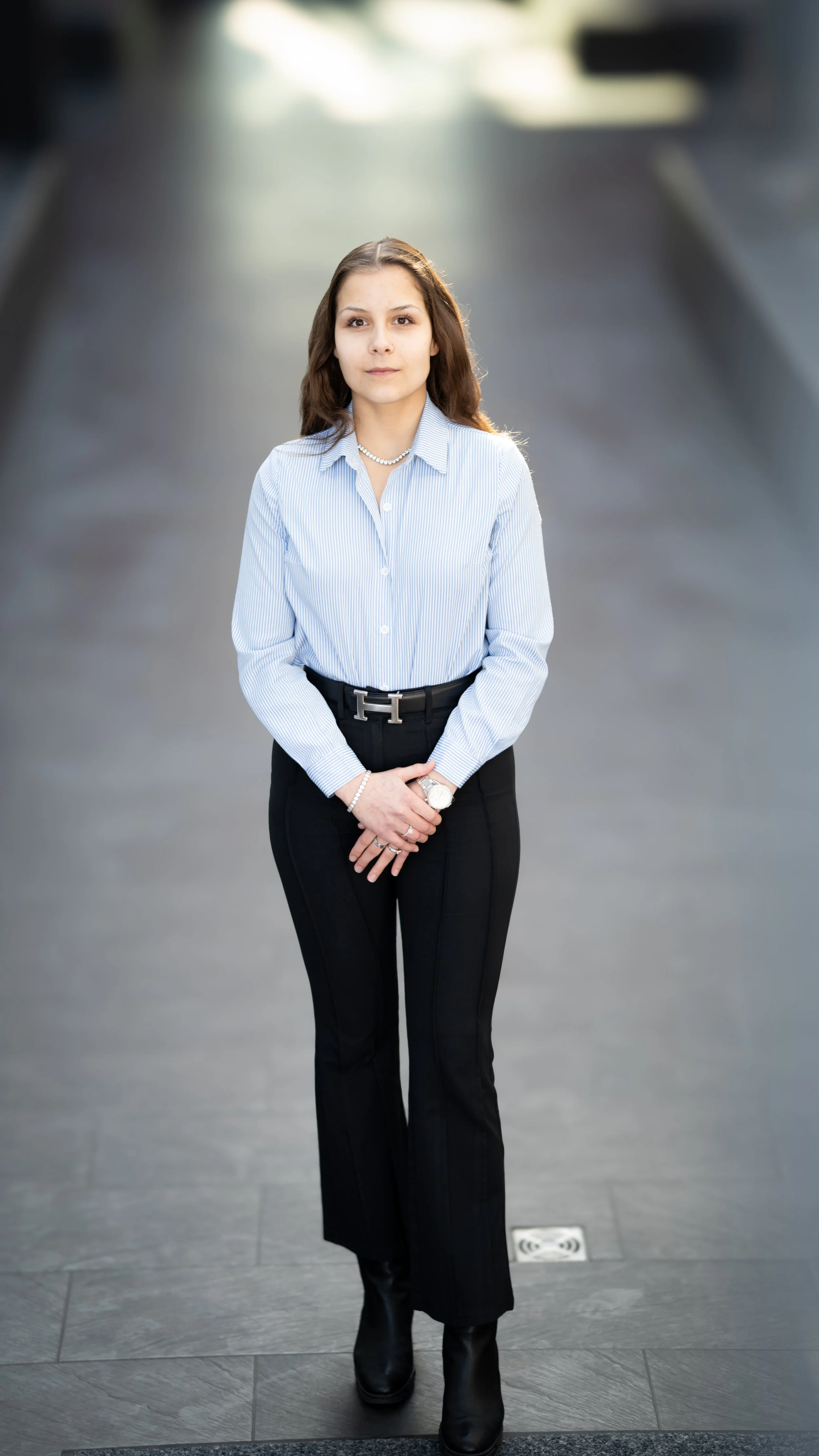 Eine junge Frau in Businesskleidung, gestylt mit einem blauen Hemd, schwarzen Hosen, schwarzen Stiefeln und Schmuck, steht auf einem Gehweg, im Hintergrund unscharf.