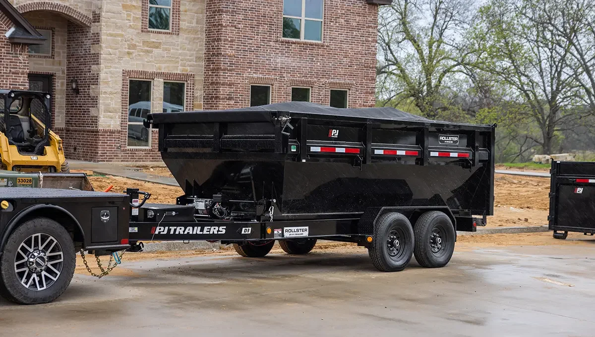 A black dump trailer hitched to a black trailer hitch, parked on a concrete surface near a brick building under construction, with construction equipment nearby and trees in the background.