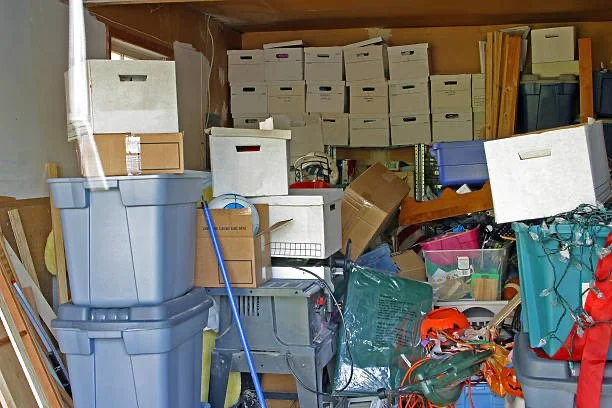 Cluttered storage room filled with boxes, plastic containers, and miscellaneous items with a wooden shelf in the background.