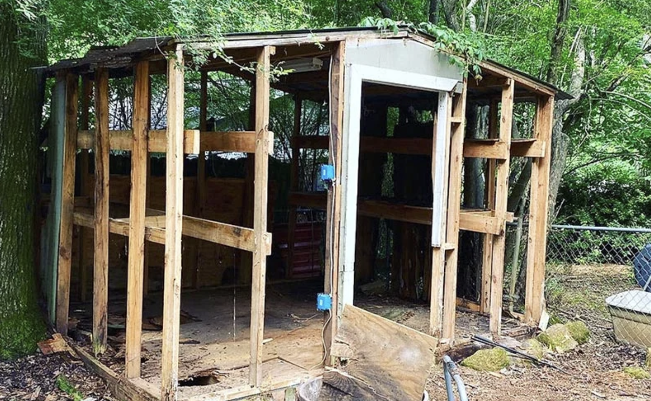 A partially built wooden structure outdoors, surrounded by trees and a chain-link fence, with construction tools and debris around.