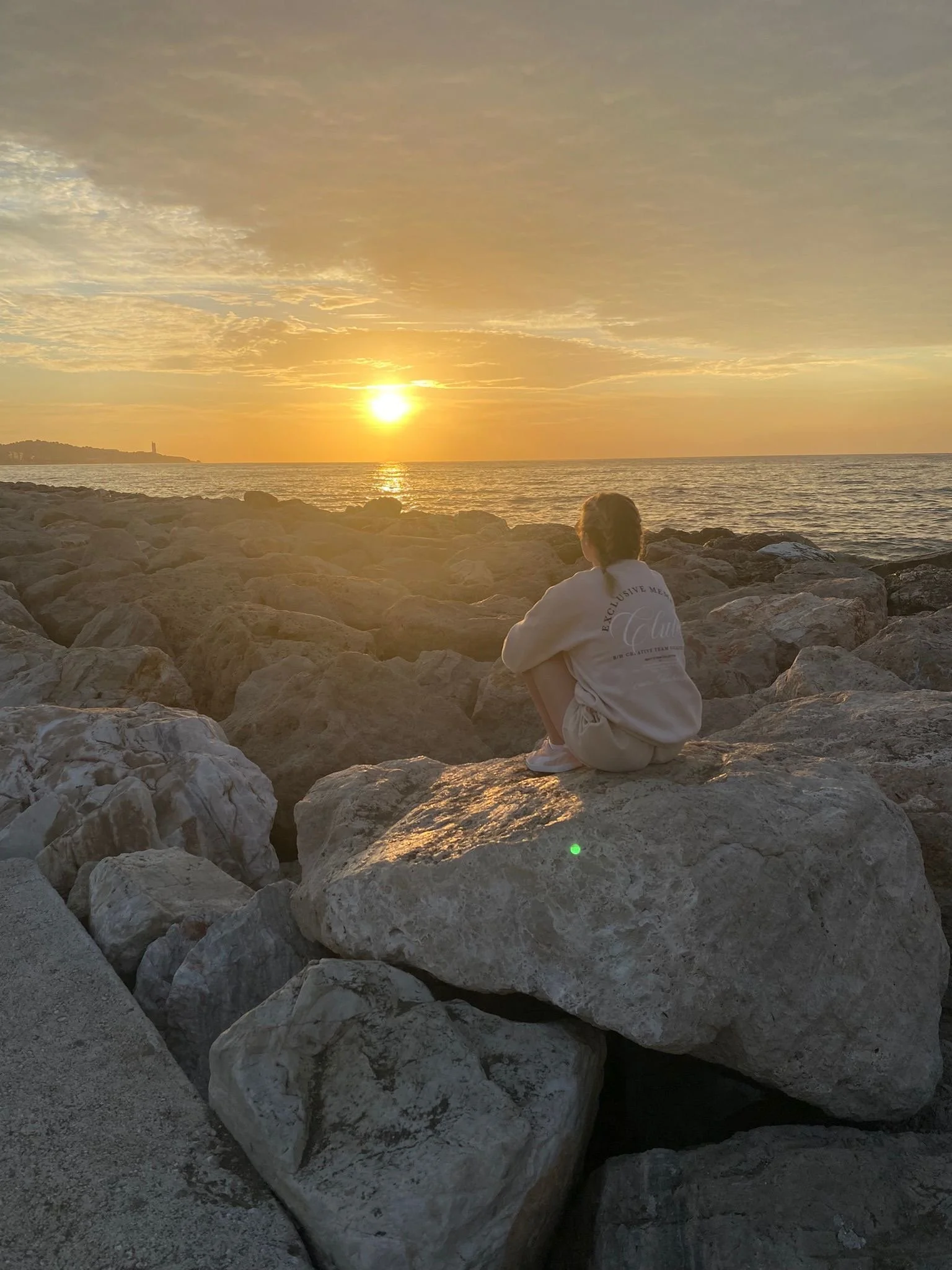Elaine doyle irish graphic designer sitting on large rocks by the beach, watching the sunset over the ocean with a cloudy sky.