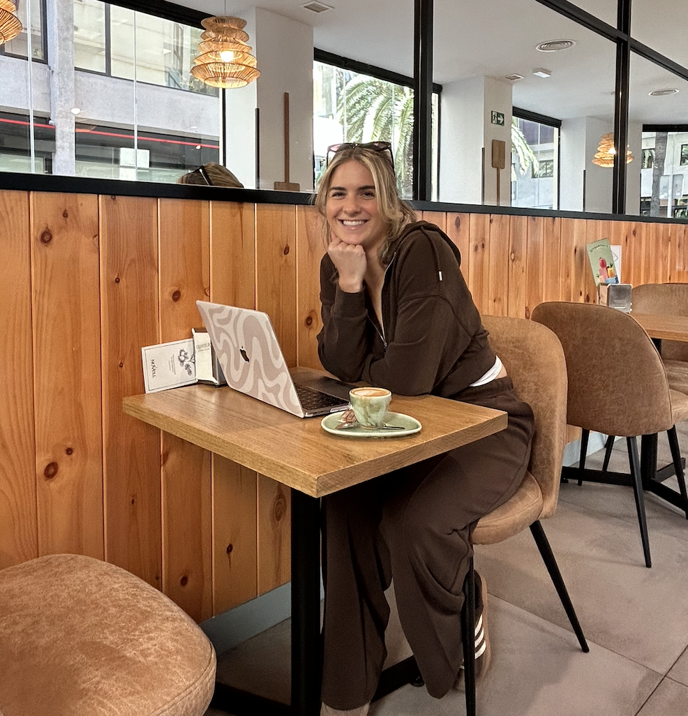 A woman with blonde hair smiling and resting her chin on her hand sits at a wooden table in a cafe, with a laptop and a cup of coffee in front of her. The cafe has large windows, wooden wall paneling, and cozy seating.