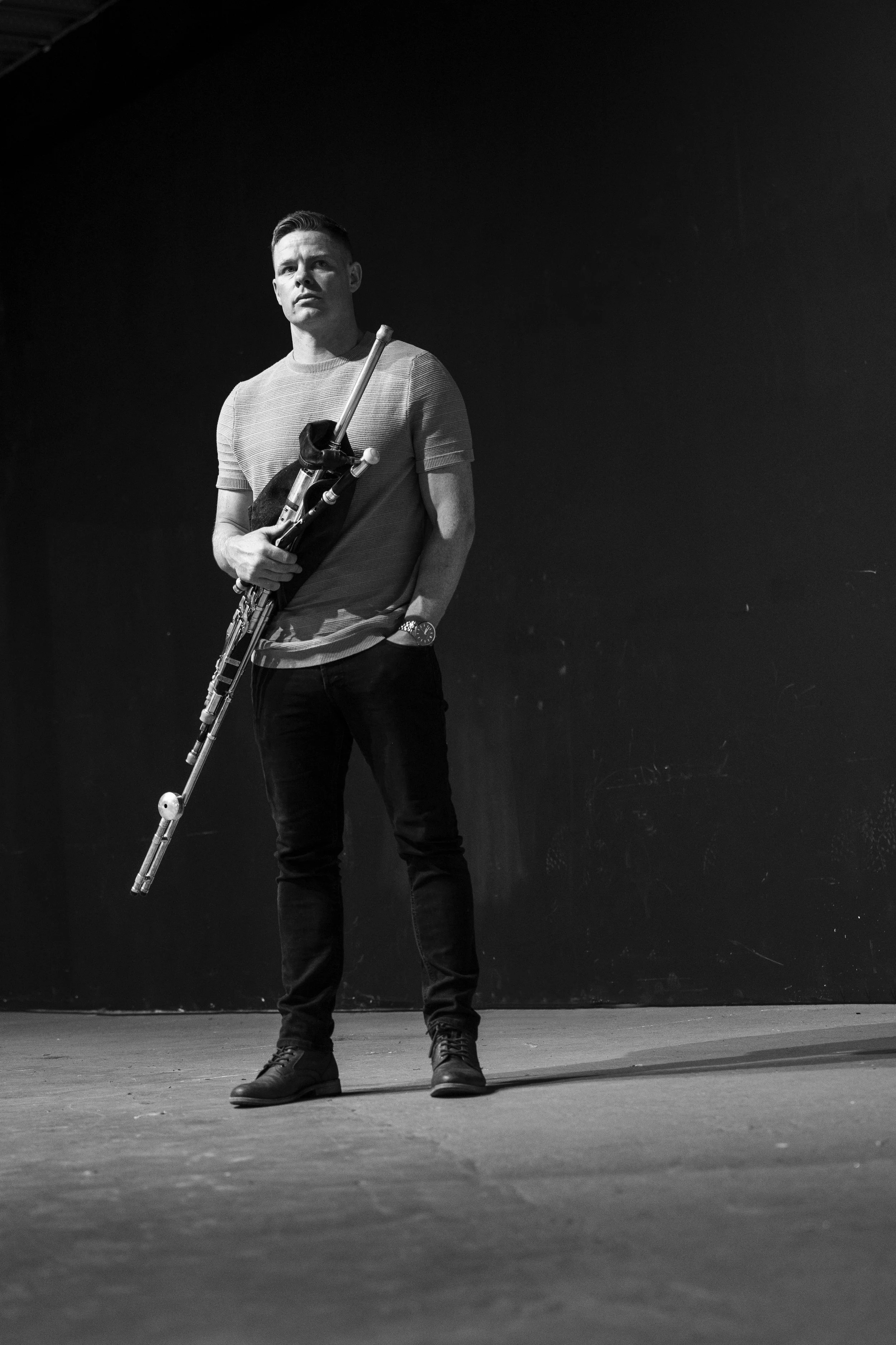 A young man standing against a dark wall holding a bassoon, dressed casually in a short-sleeved shirt, dark pants, and boots, with a watch on his left wrist.