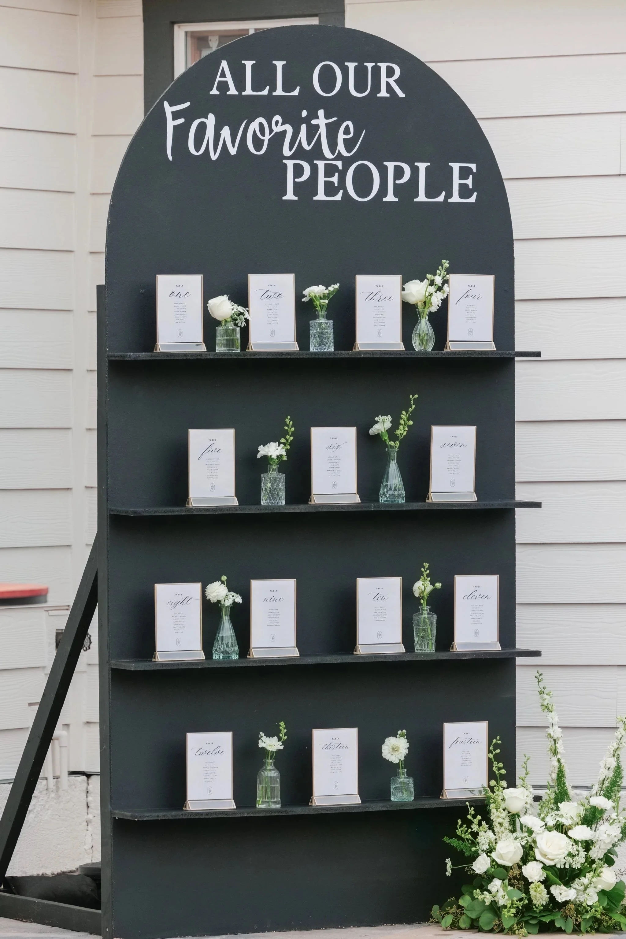 A decorative display with a black stand and shelves holding framed table numbers and small vases with white flowers, positioned against a beige wall. The top of the stand reads, 'ALL OUR Favorite PEOPLE'.