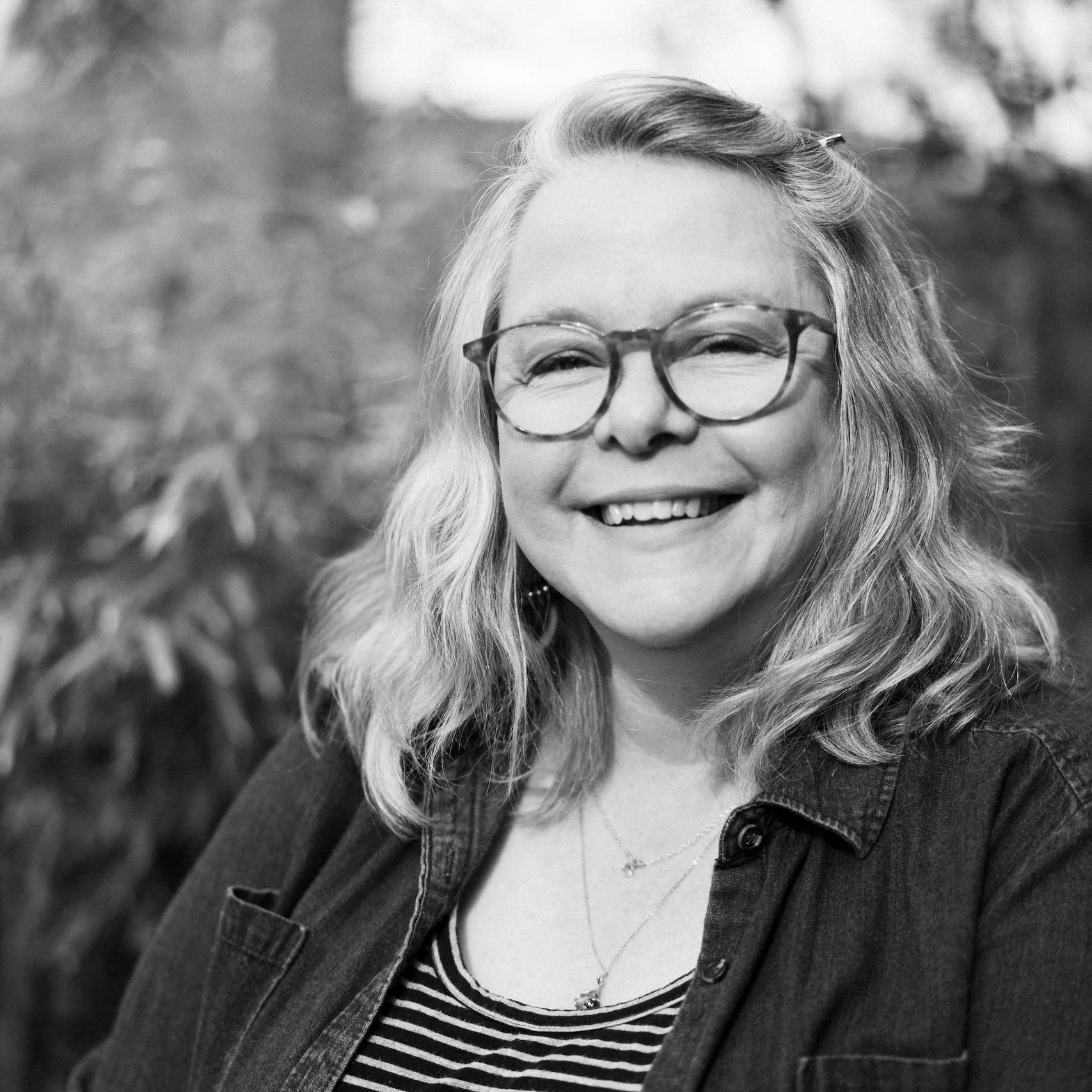 A woman with wavy shoulder-length hair, wearing glasses, a striped shirt, and a denim jacket, smiling in an outdoor setting.