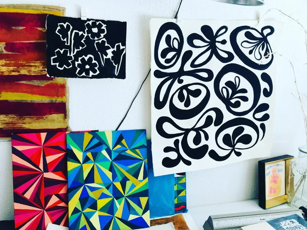 Various colorful abstract paintings and black-and-white artwork displayed on a wall and desk area, including floral and geometric designs.