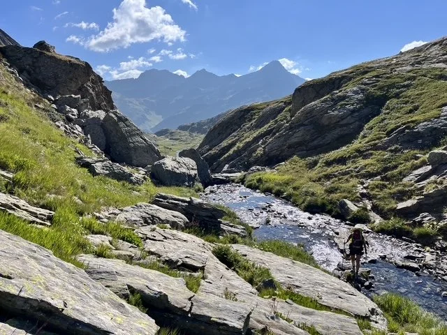 Ein Wanderer entlang eines Flusses in den Bergen, umgeben von grünen Hängen und steinigen Pfaden, mit hohen Gipfeln im Hintergrund bei Sonnenschein.