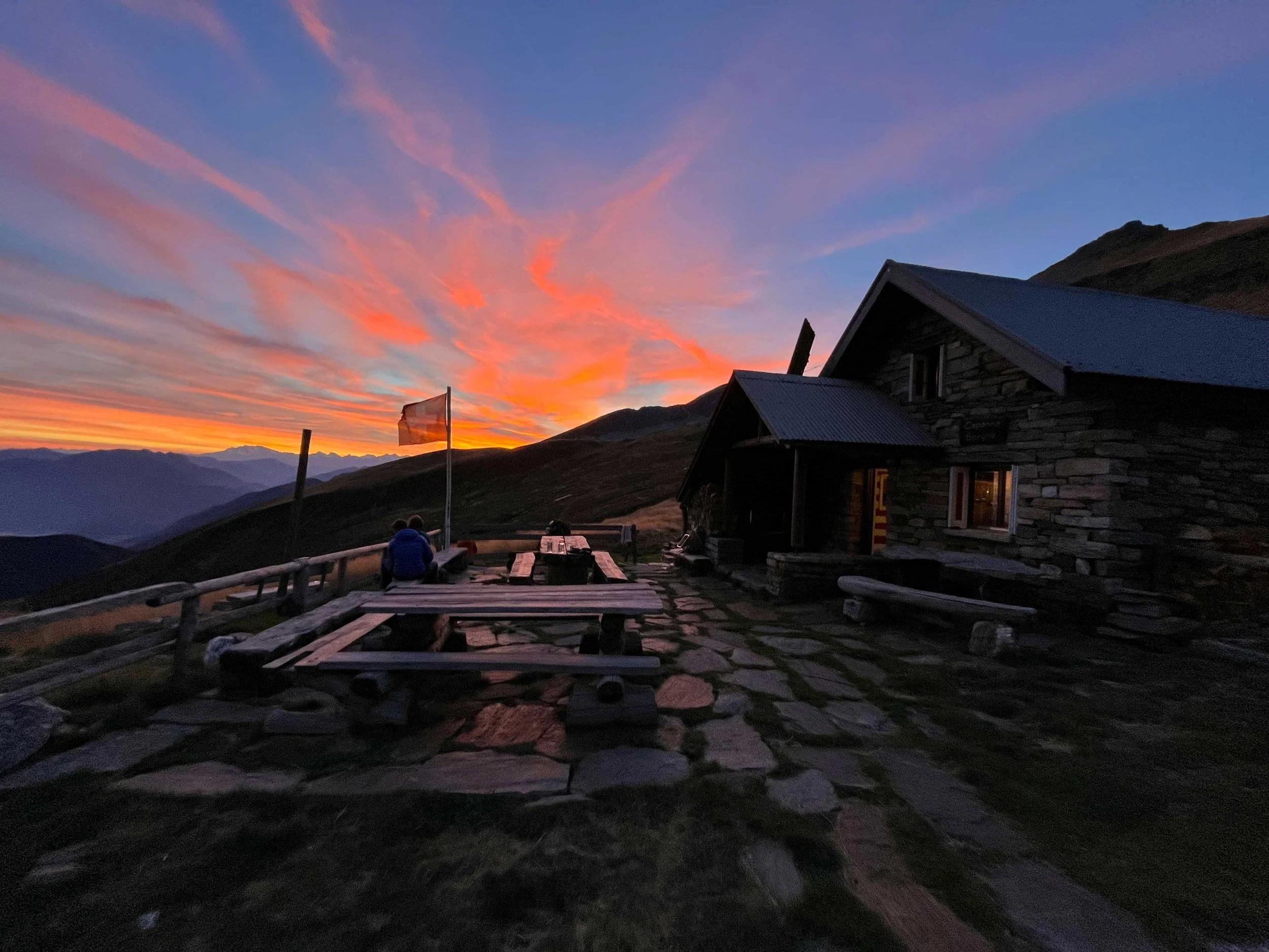 Ein abgelegenes Berghütte im Sonnenuntergang mit einem Holz-Tisch und Bänken im Vordergrund, umgeben von Bergen bei Dämmerung.