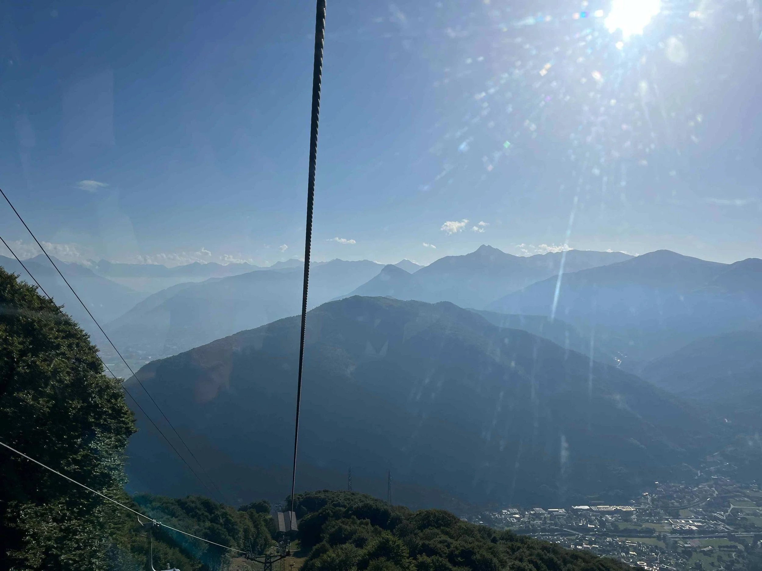 Berglandschaft mit einer Seilbahn und Kabeln im Vordergrund, Himmel mit Sonne und wenigen Wolken im Hintergrund.