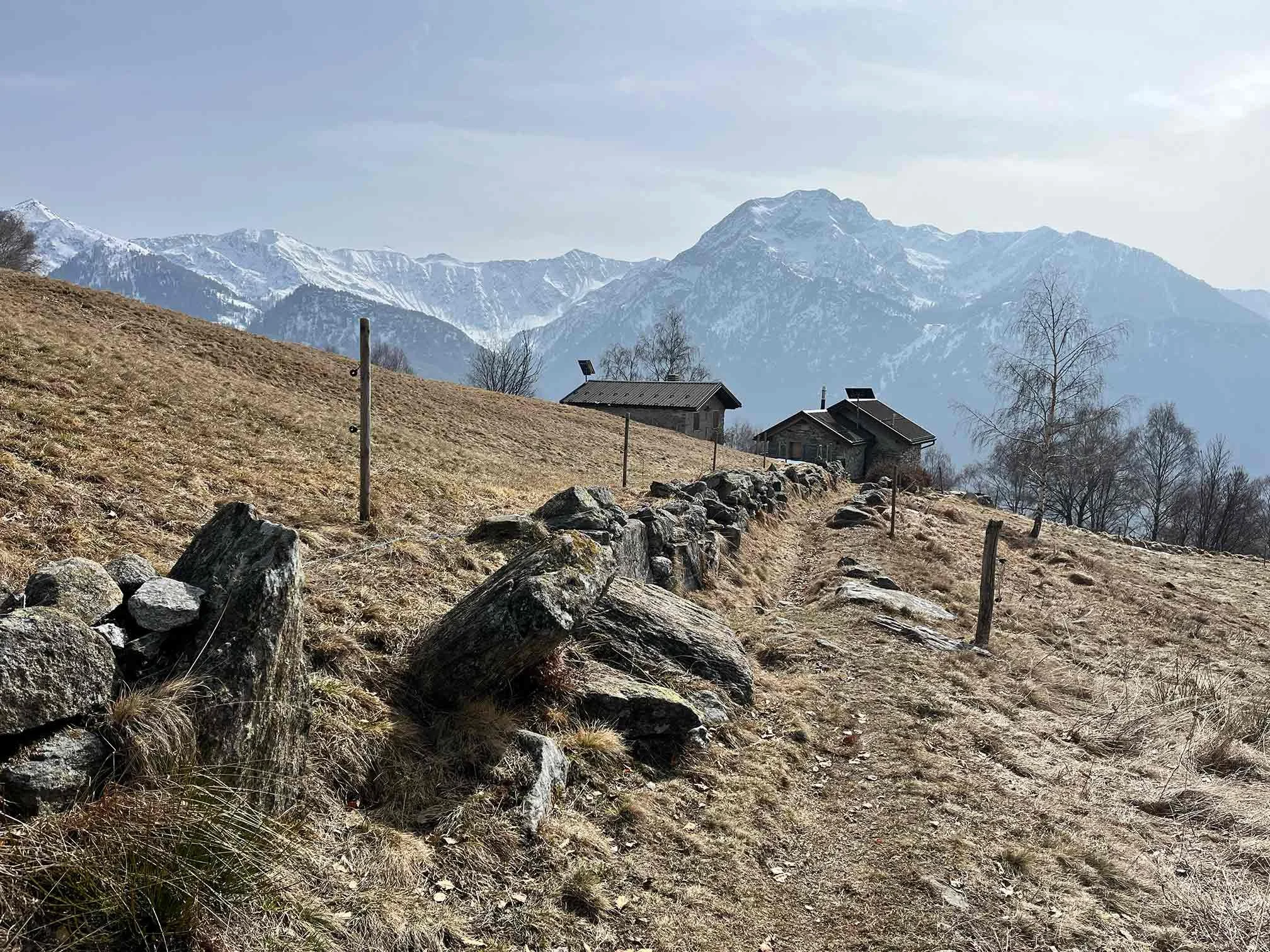 Wanderweg, wenige Häuser und mit Schnee bedeckte Berge im Hintergrund, karg, trockenes Gras.