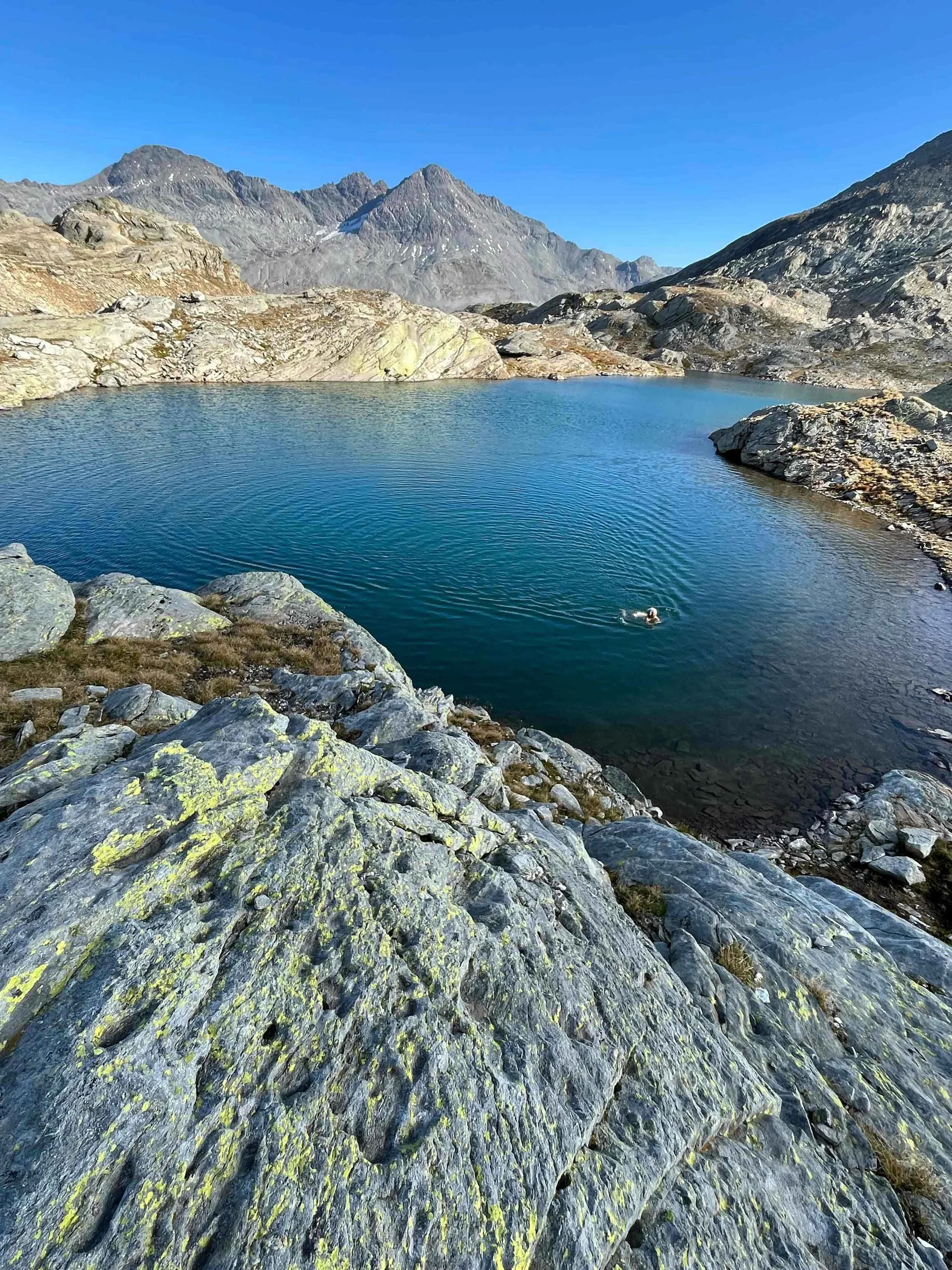 Junge Frau mit Rucksack am See in den Bergen, bewölkter Himmel, grüne Wiesen und Berge im Hintergrund.