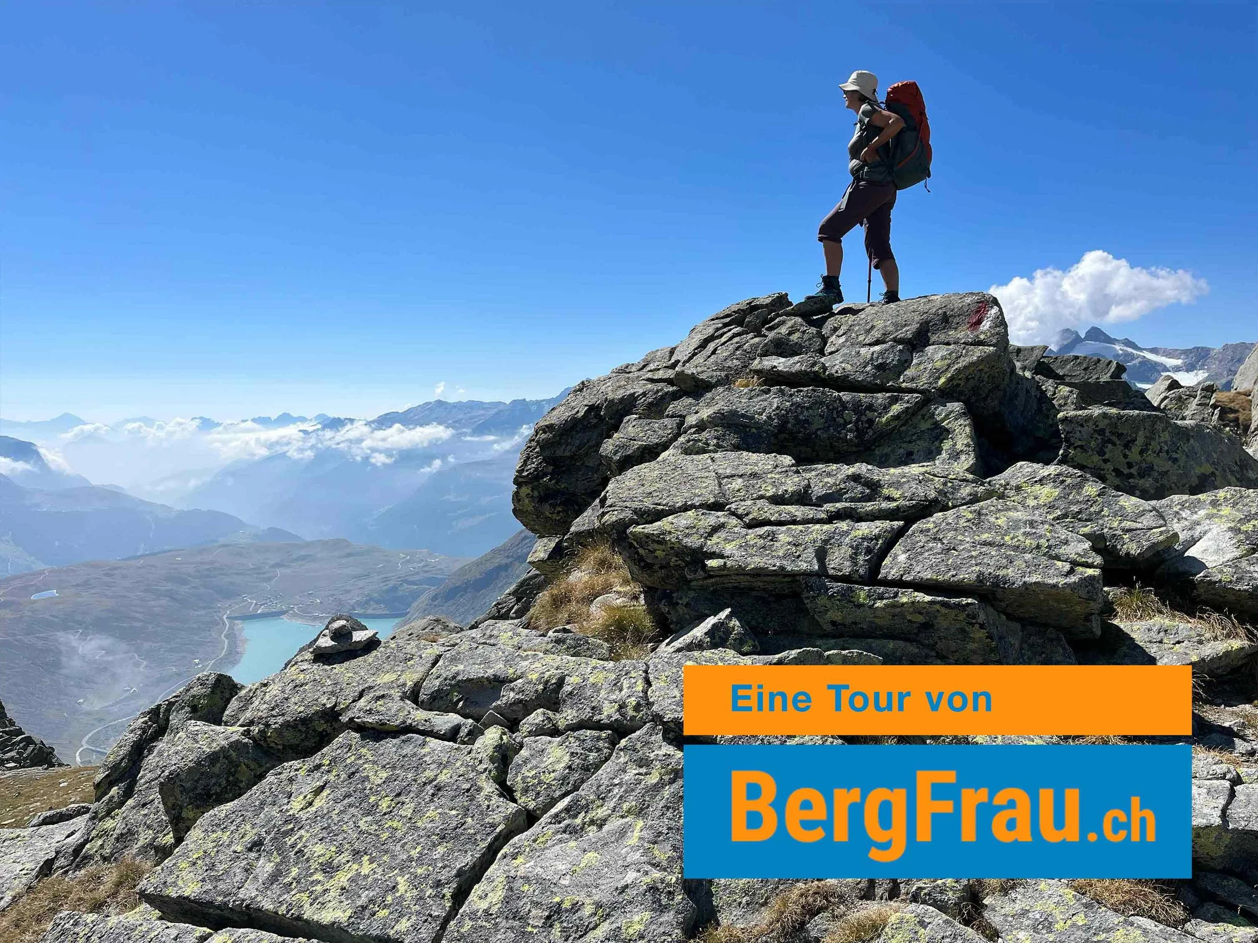 Ein Bergsteiger mit Rucksack und Hut steht auf einem felsigen Gipfel unter blauem Himmel mit Wolken im Hintergrund.