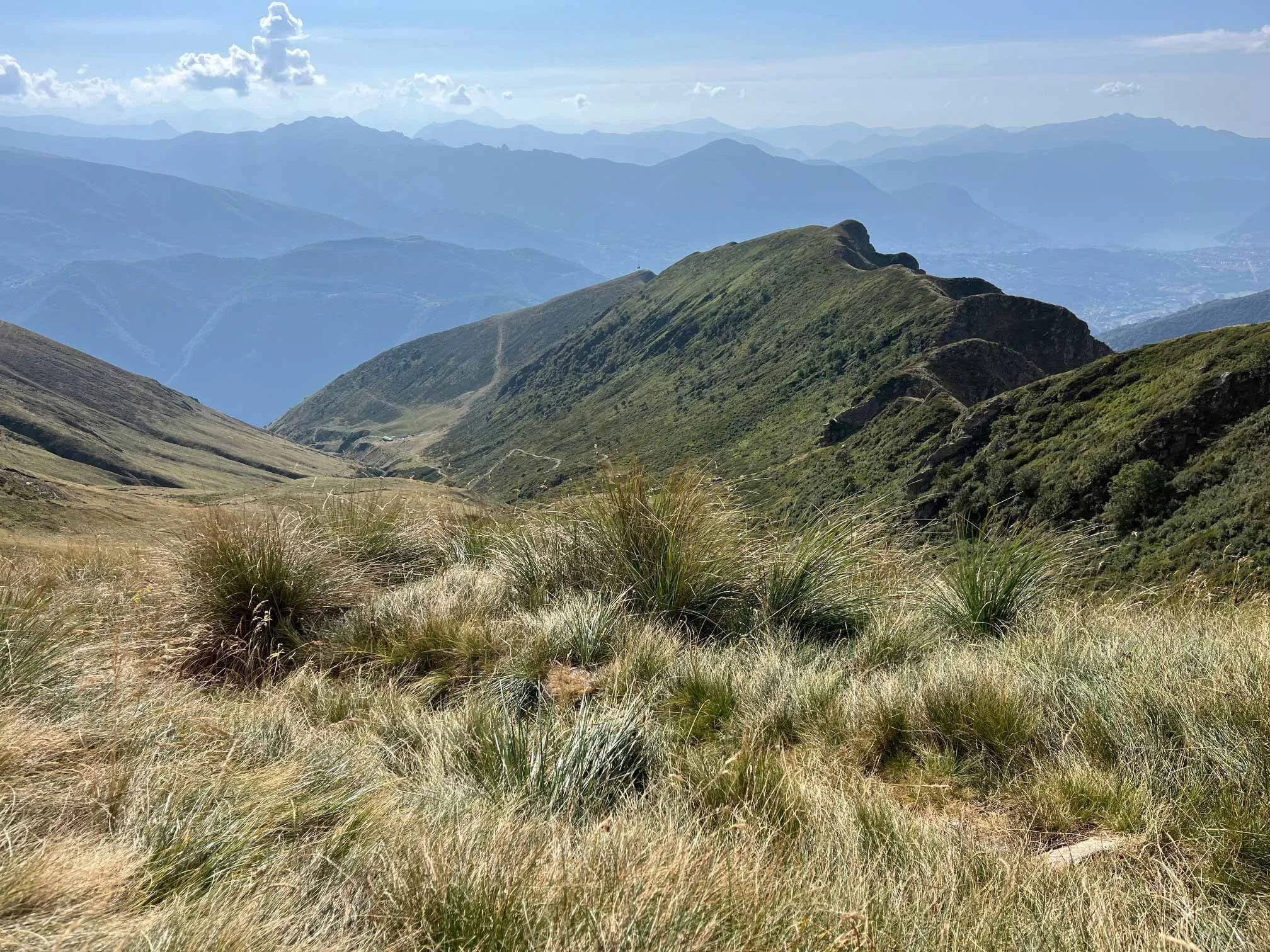 Berglandschaft mit grünen Bergen und Hügeln, im Hintergrund sind weitere Berge und eine bewölkte Himmelsszene zu sehen.