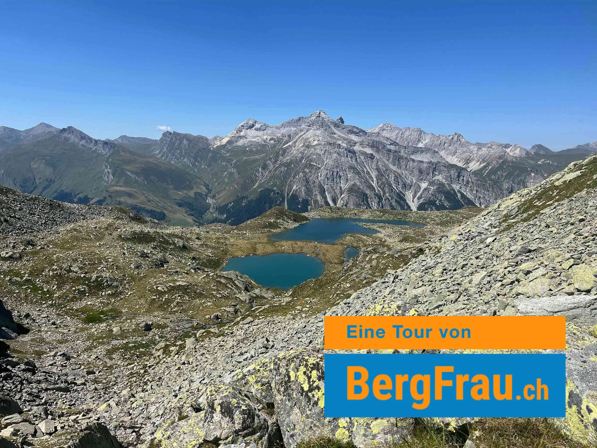 Bergsee in den Alpen mit Bergen im Hintergrund, blauer Himmel, Steine und Gras im Vordergrund. Text auf dem Bild: ‚Eine Tour von BergFrau.ch‘.